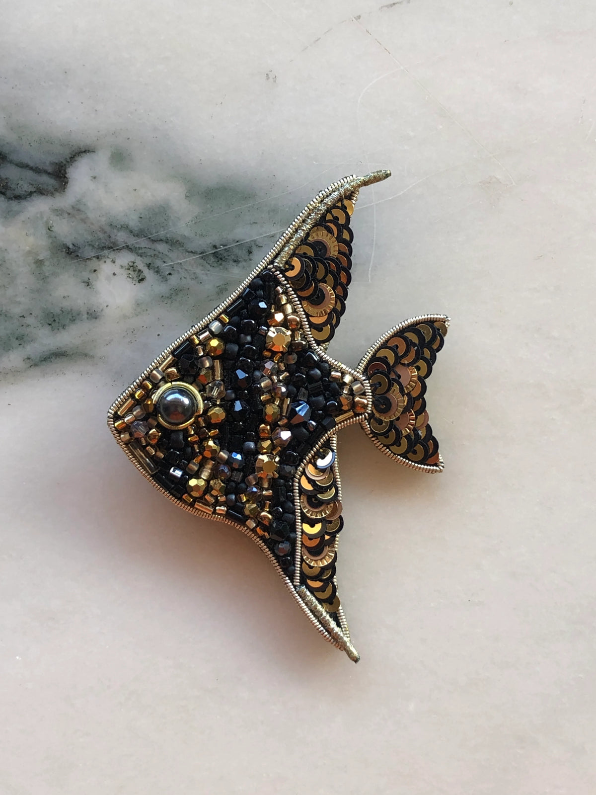 Broochella Men’s Fish Brooch Pin featuring black and gold beadwork, metallic sequins and crystal embellishments, photographed on a white marble background with grey-green veining.