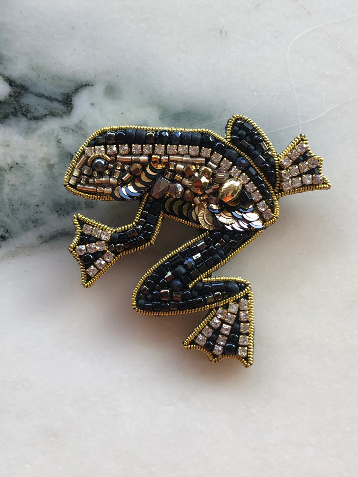 Broochella Men’s Frog Brooch Pin featuring intricate black and gold beadwork, sequins and crystal embellishments, photographed on a white marble background with grey-green veining.
