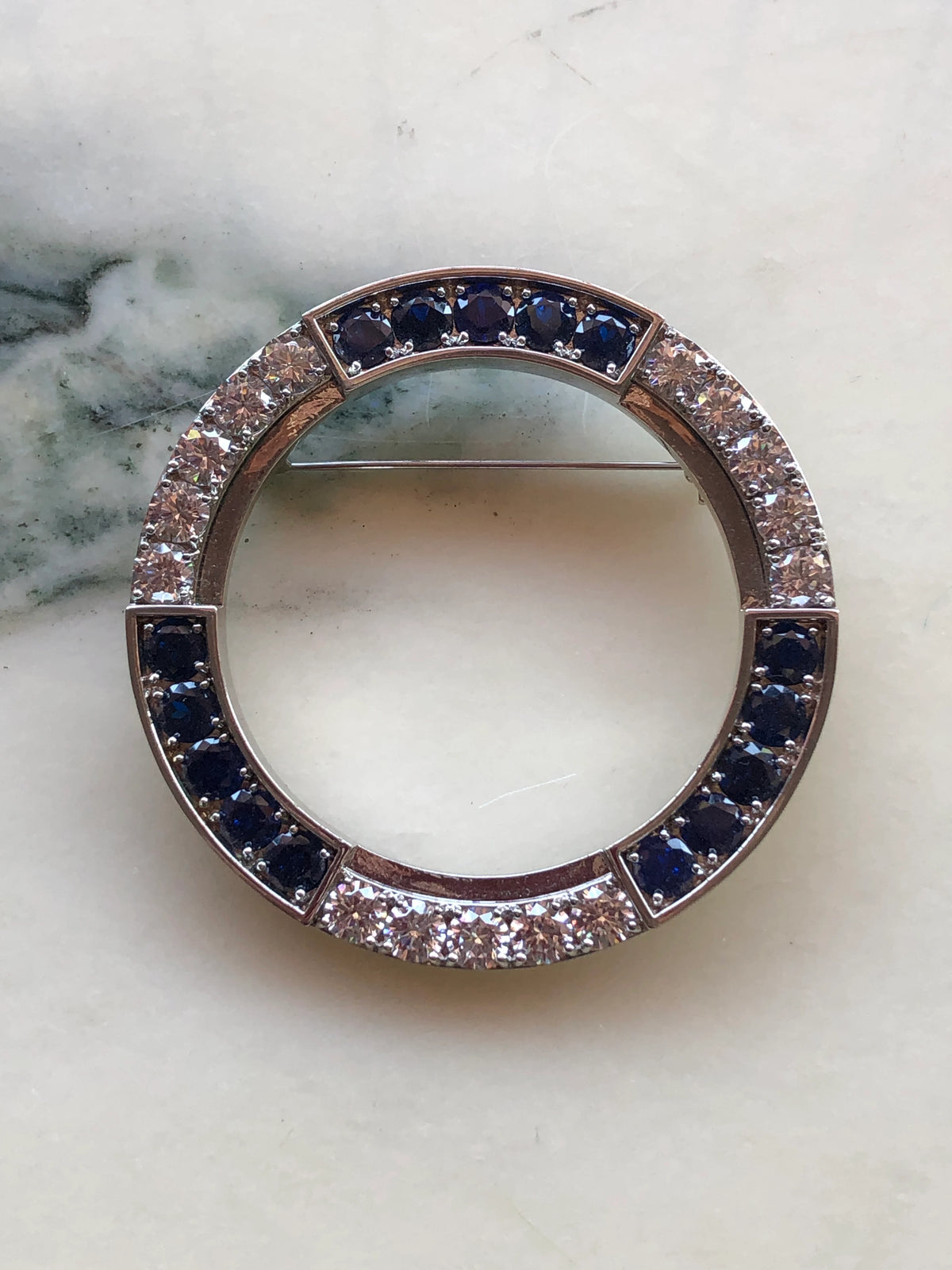 Broochella Men’s Full Moon Brooch featuring a circular silver-tone design set with deep navy blue and sparkling clear crystals, photographed on a pale white marble background with grey veining.