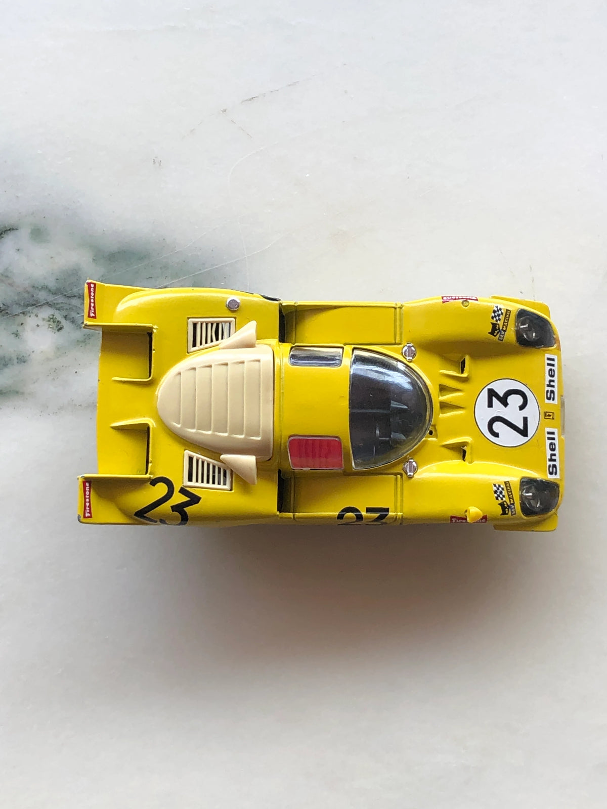 Broochella Men's Giallo Sprint No.23 Vintage Endurance Racing Car Brooch viewed from above, showcasing its yellow body, black number 23 racing decals, enclosed cockpit, cream engine cover, rear spoiler and Shell and Firestone branding.