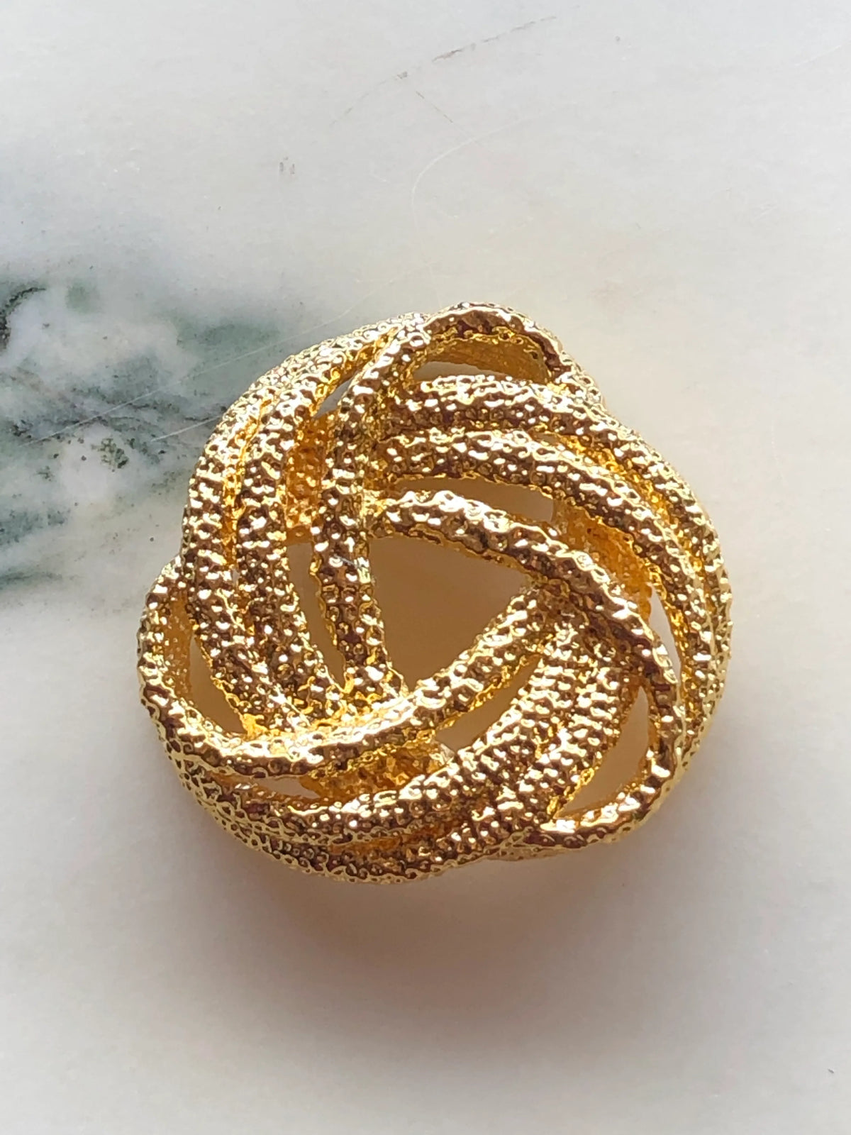 Broochella Men’s Golden Knot Brooch Pin, a sculptural designer brooch with intertwined textured gold loops forming an intricate statement knot, photographed on white marble.