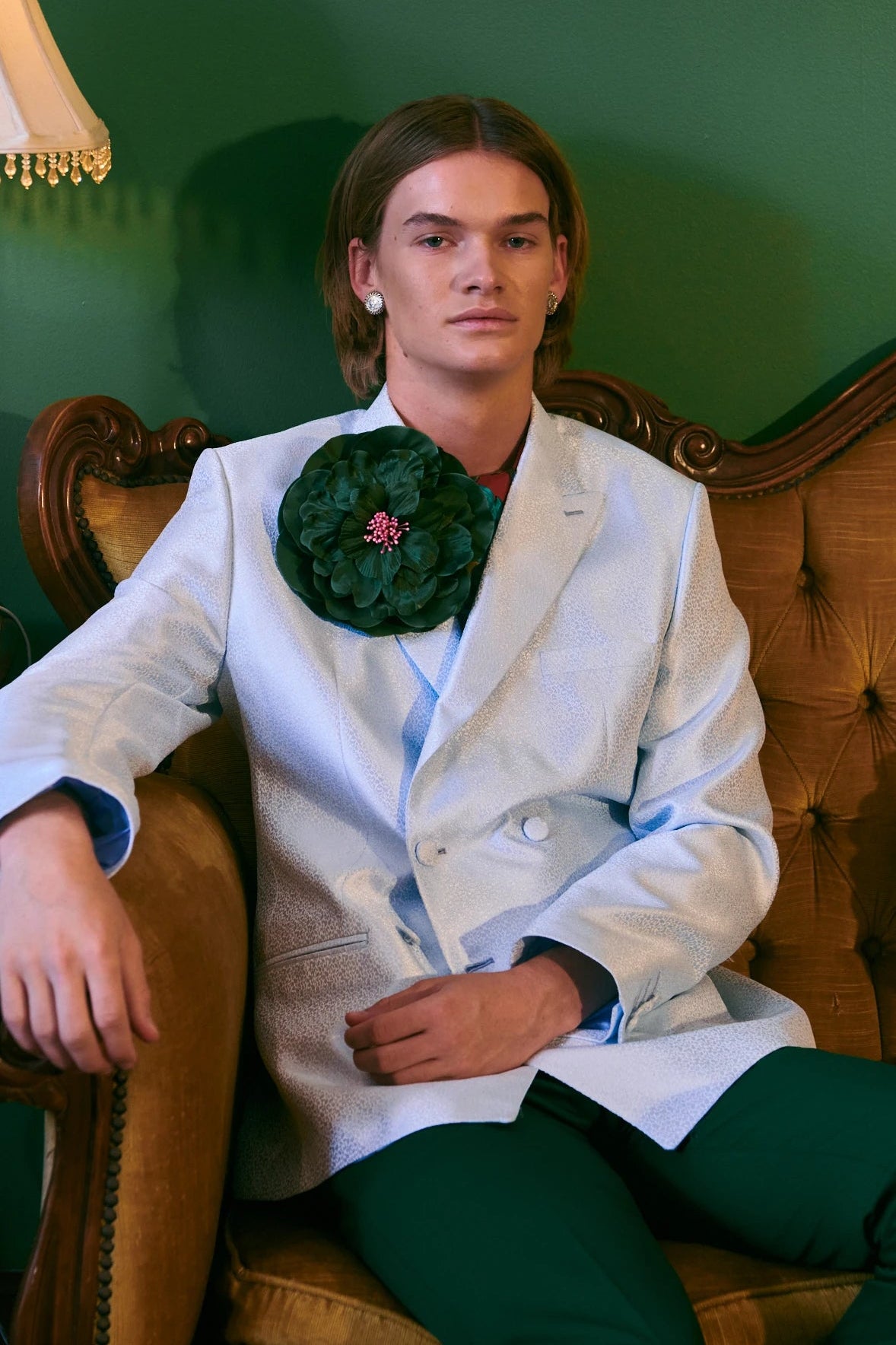 A man wearing a white double-breasted blazer with a Broochella Ibisco handmade, large dark green, fabric flower brooch, paired with dark green trousers, sitting on a vintage tufted brown armchair against a dark green wall.