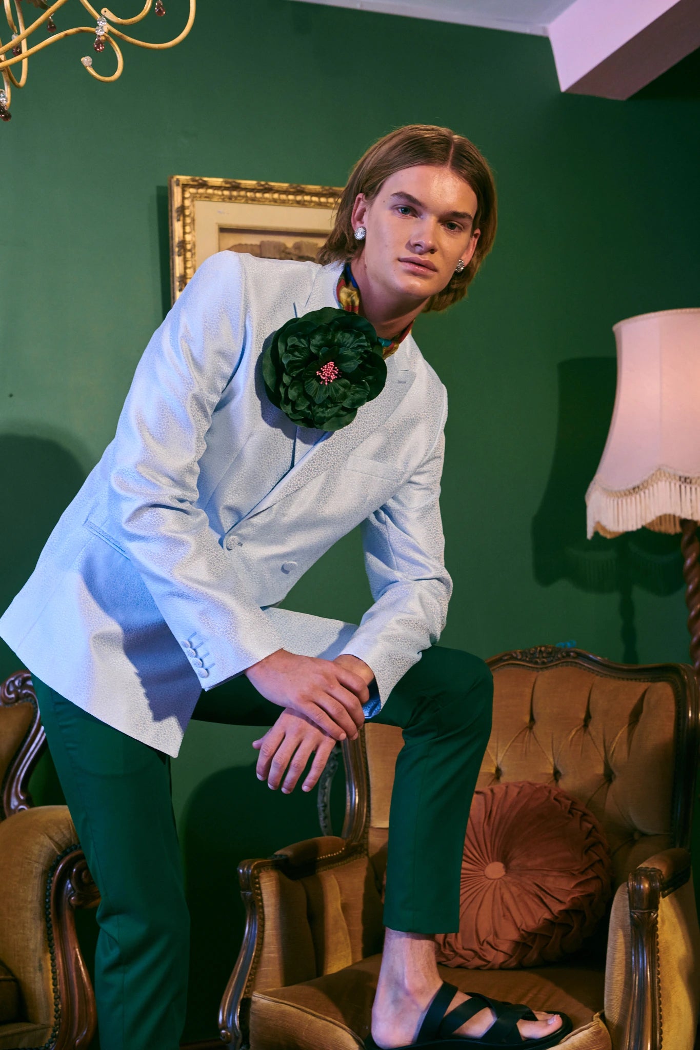 A man wearing a white double-breasted blazer with a Broochella Ibisco handmade, large dark green, fabric flower brooch, paired with dark green trousers, sitting on a vintage tufted brown armchair against a dark green wall.