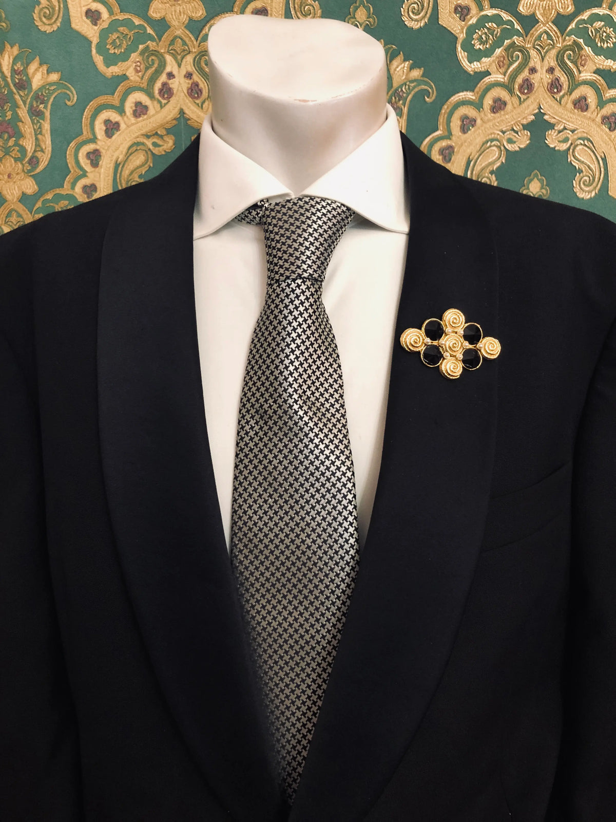 Broochella Men's Isabella vintage black and gold statement brooch featuring ornate gold-tone spiral detailing and four faceted black stones in a geometric design, styled on the lapel of a black tailored suit with a white shirt and black-and-white houndstooth tie, against emerald green and gold damask wallpaper.