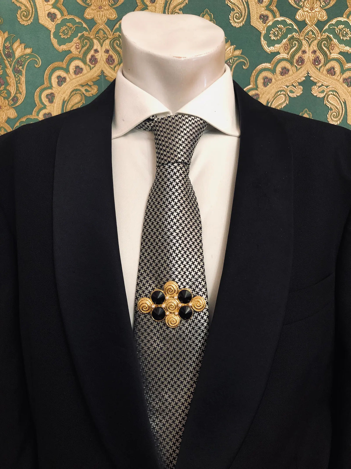 Broochella Men's Isabella vintage black and gold brooch featuring sculptural gold-tone scrollwork, four glossy faceted black stones and decorative spiral motifs, pinned to a black-and-white houndstooth necktie with a crisp white shirt and black tailored suit, photographed against ornate emerald green and gold damask wallpaper.