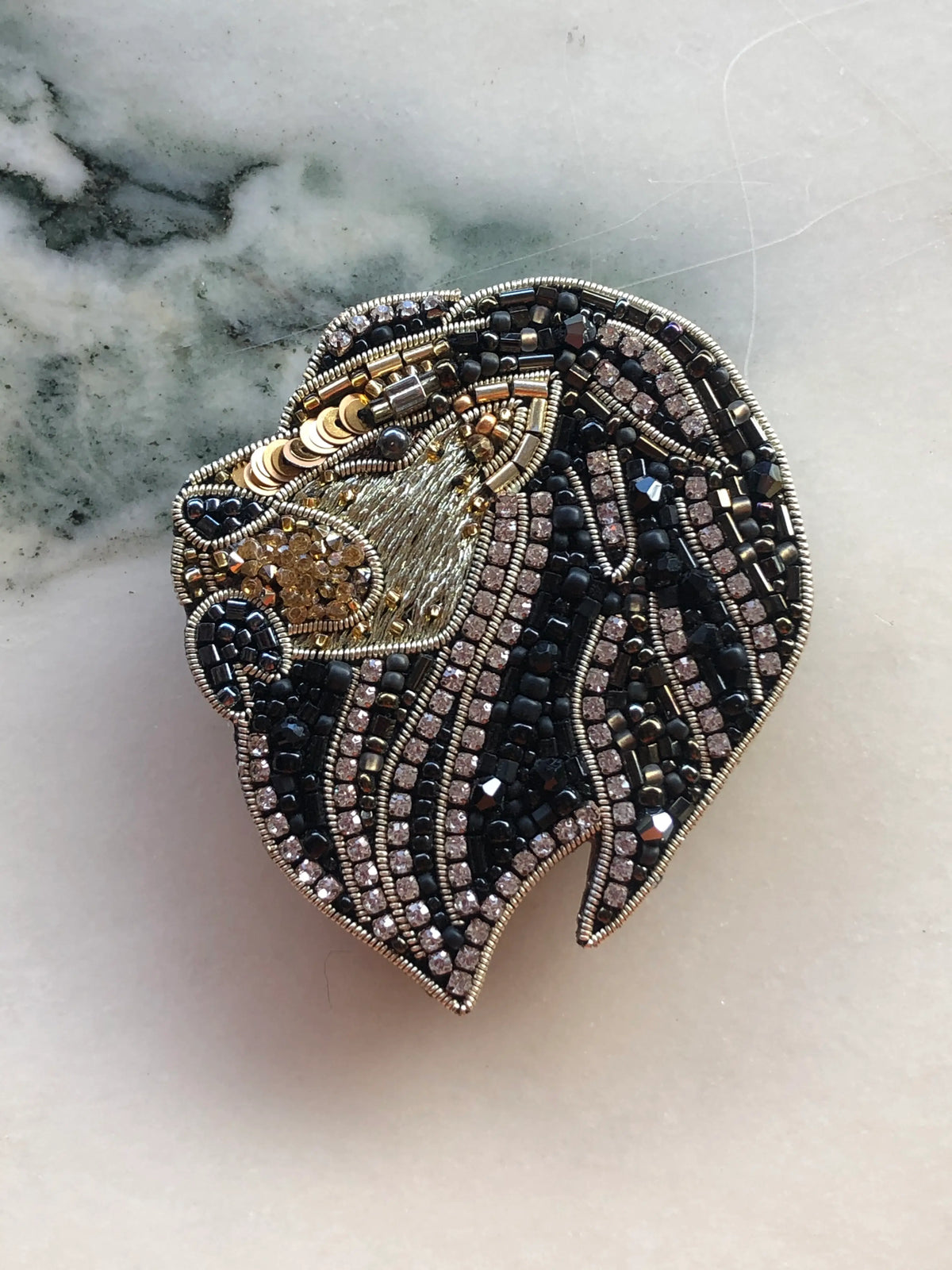Broochella Men’s Lion Brooch Pin featuring an intricately beaded lion head in black, gold and silver tones with crystal detailing and a sweeping embellished mane, photographed on a white marble background with grey-green veining.