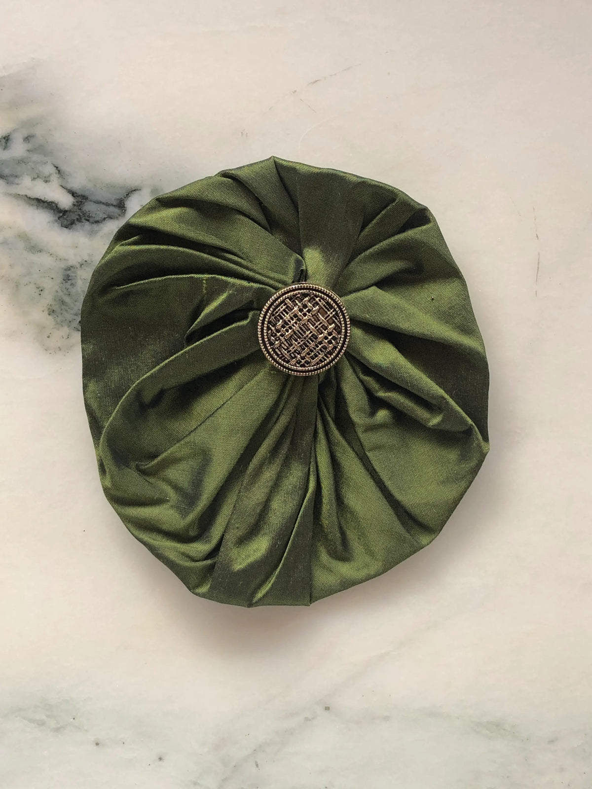 Broochella Men’s Lollipop Brooch in olive green, large size, featuring voluminous gathered silk fabric with an antique-style metal centre, photographed on a pale white marble background with grey-green veining.