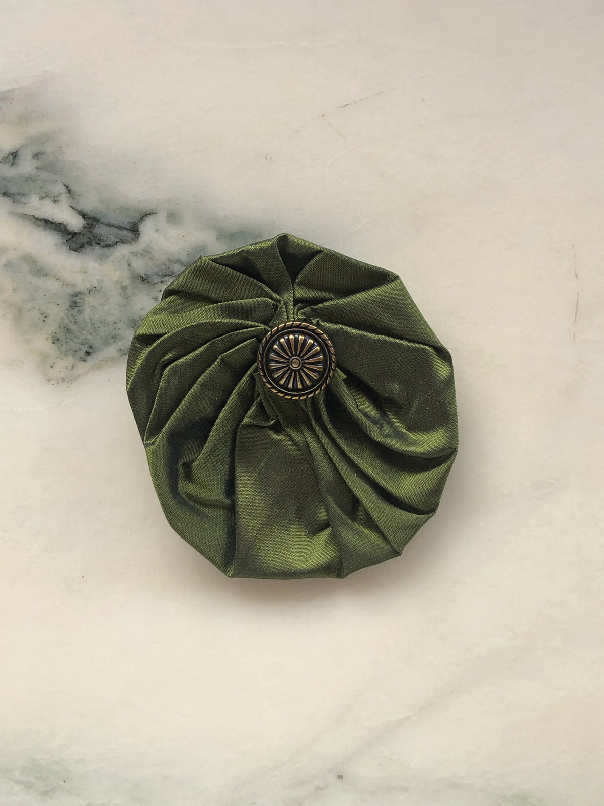 Broochella Men’s Lollipop Brooch in olive green, medium size, featuring gathered silk fabric with an antique-style metal centre, photographed on a pale white marble background with grey-green veining.