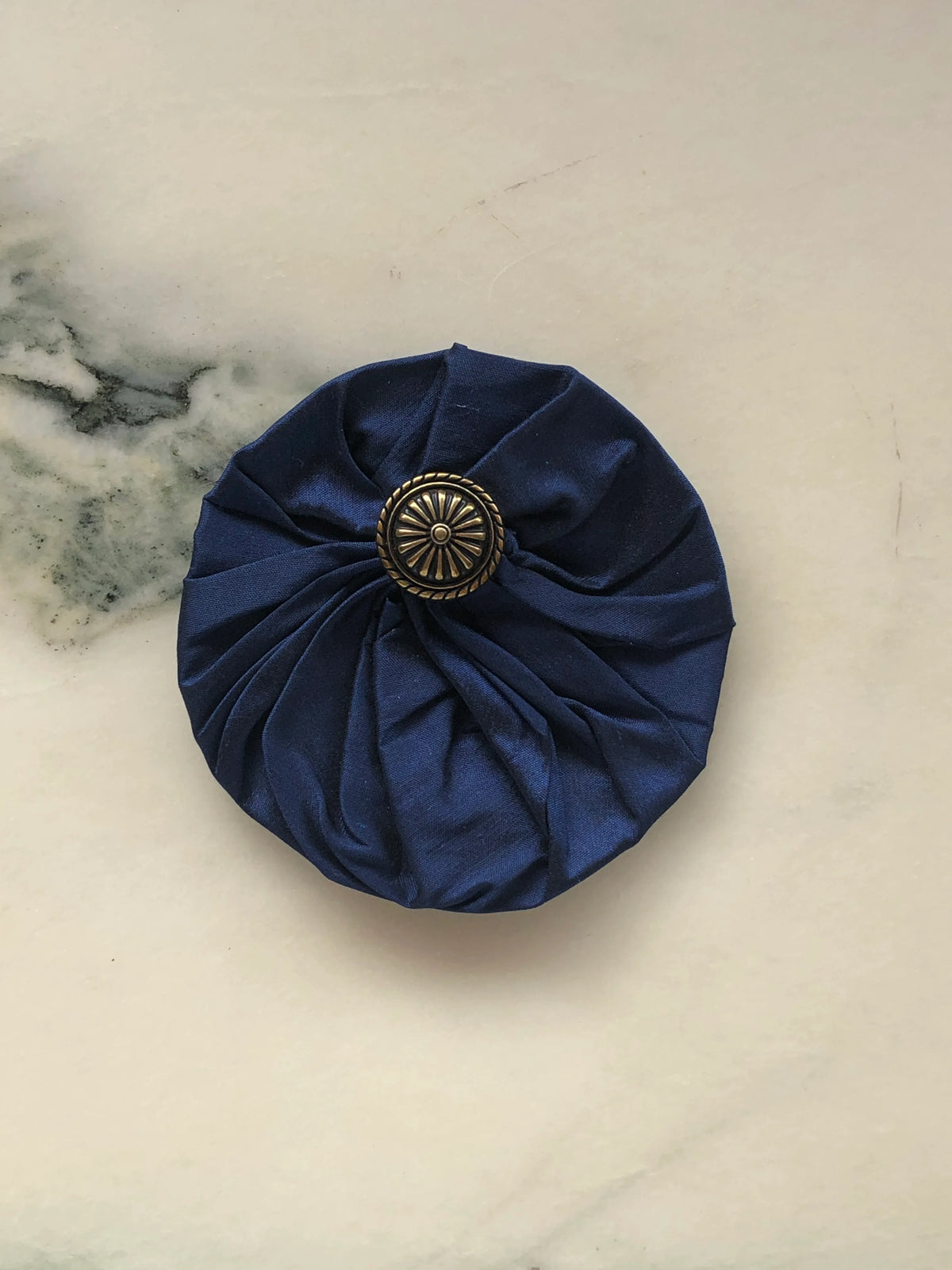 Broochella Men’s Lollipop Brooch in navy, small size, featuring gathered silk fabric with a decorative silver-tone centre, photographed on a pale white marble background with grey-green veining.