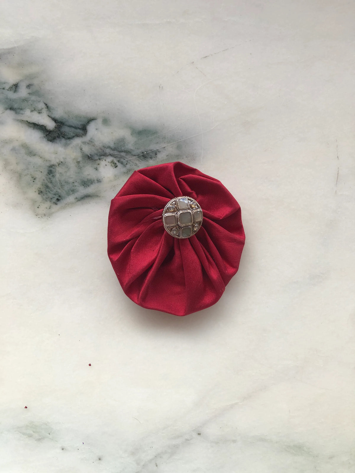 Broochella Men’s Lollipop Brooch in red, small size, featuring gathered silk fabric with a decorative silver-tone centre, photographed on a pale white marble background with grey-green veining.