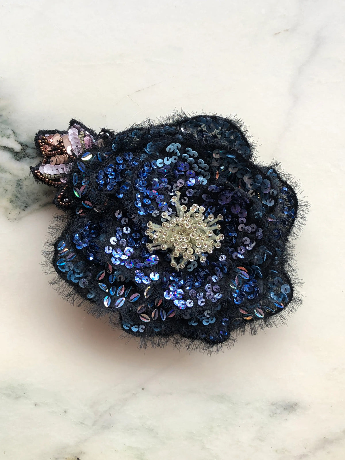 Broochella Men's Luminosa Small Flower Brooch Pin in blue, featuring layered black fabric petals embellished with shimmering navy, sapphire blue and iridescent sequins, a delicate ivory beaded centre and decorative sequinned leaves. A statement floral brooch for men's blazers, jackets and occasionwear.