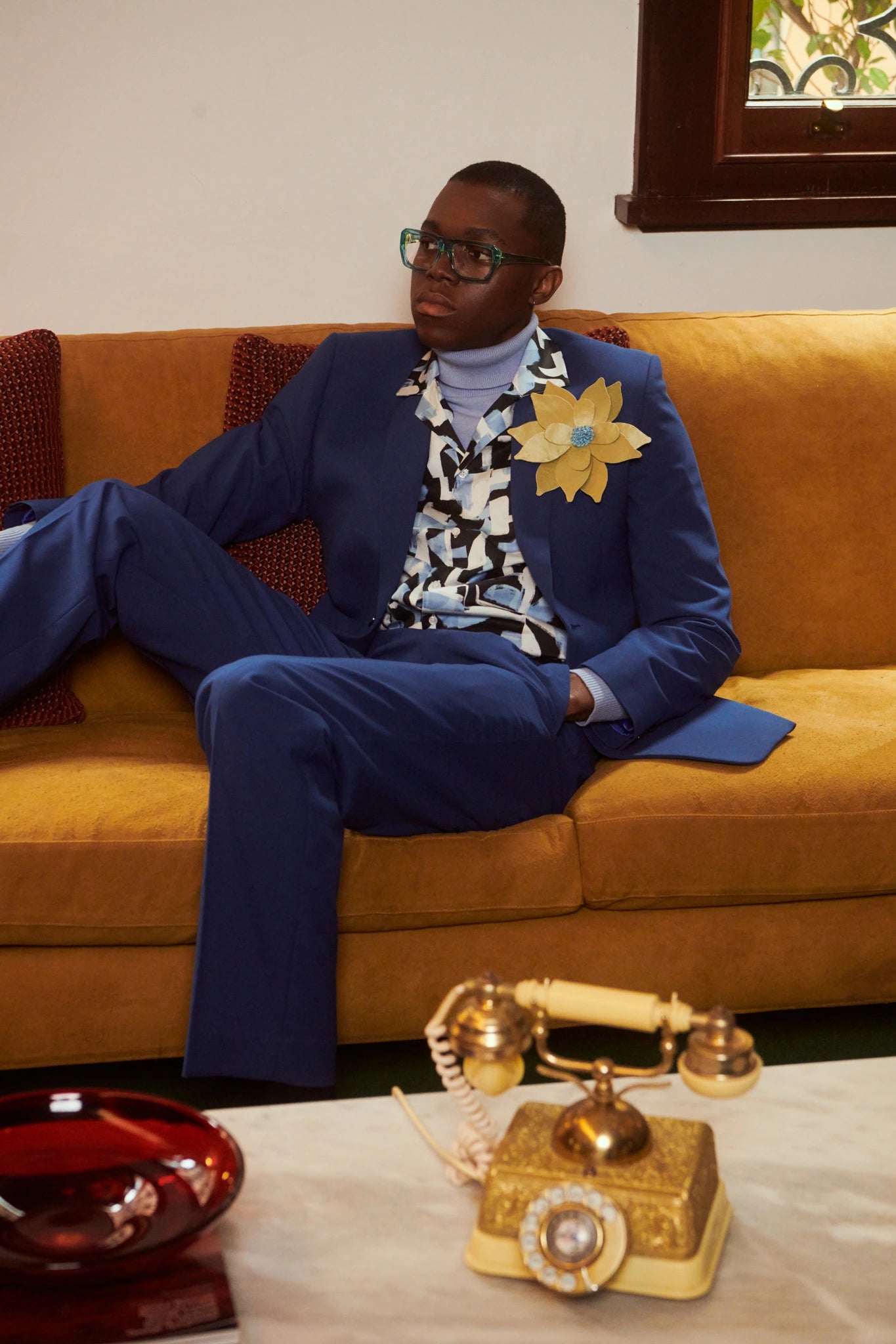 A man wearing a blue suit, patterned shirt, and Broochella ninfea green flower lapel pin sits on a mustard yellow couch in a living room setting.