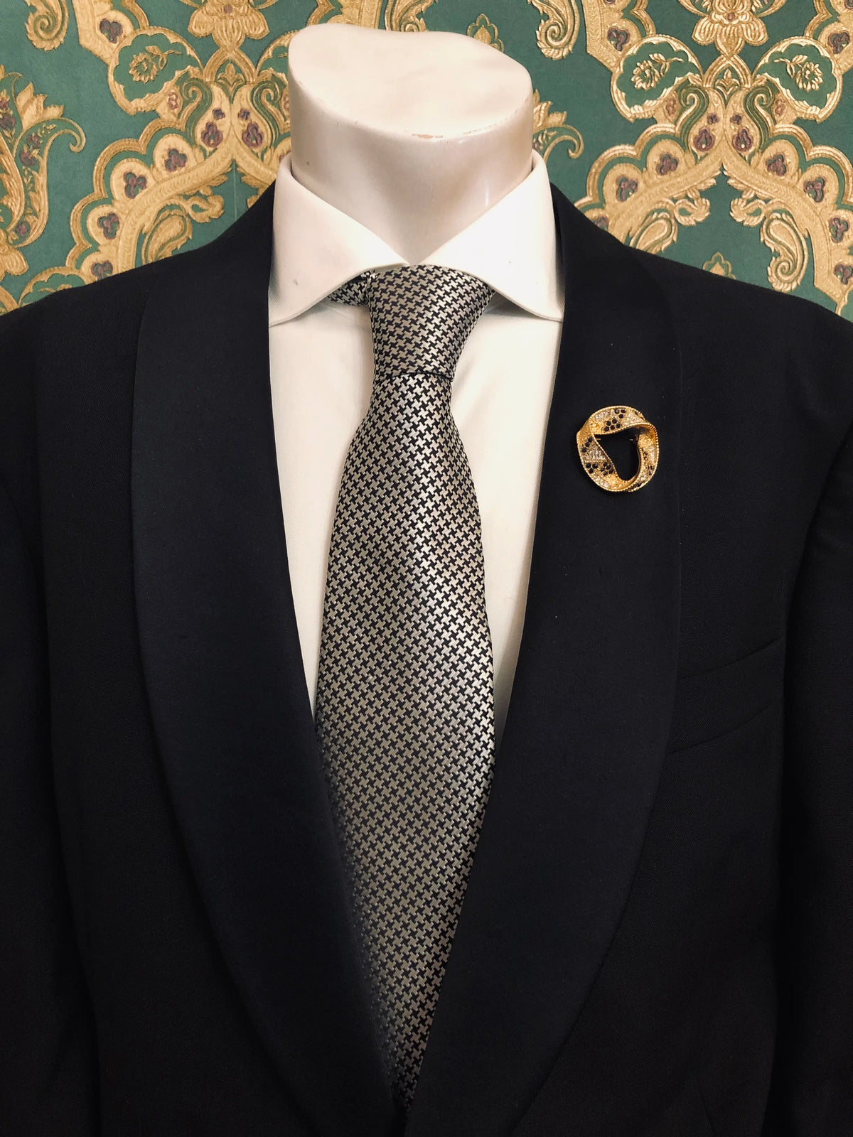 Broochella Men's Ottavia vintage gold-tone swirl brooch featuring an interlocking circular design embellished with black and clear sparkling rhinestones, styled on the lapel of a black tailored suit with a white shirt and black-and-white houndstooth tie, against ornate emerald green and gold damask wallpaper.