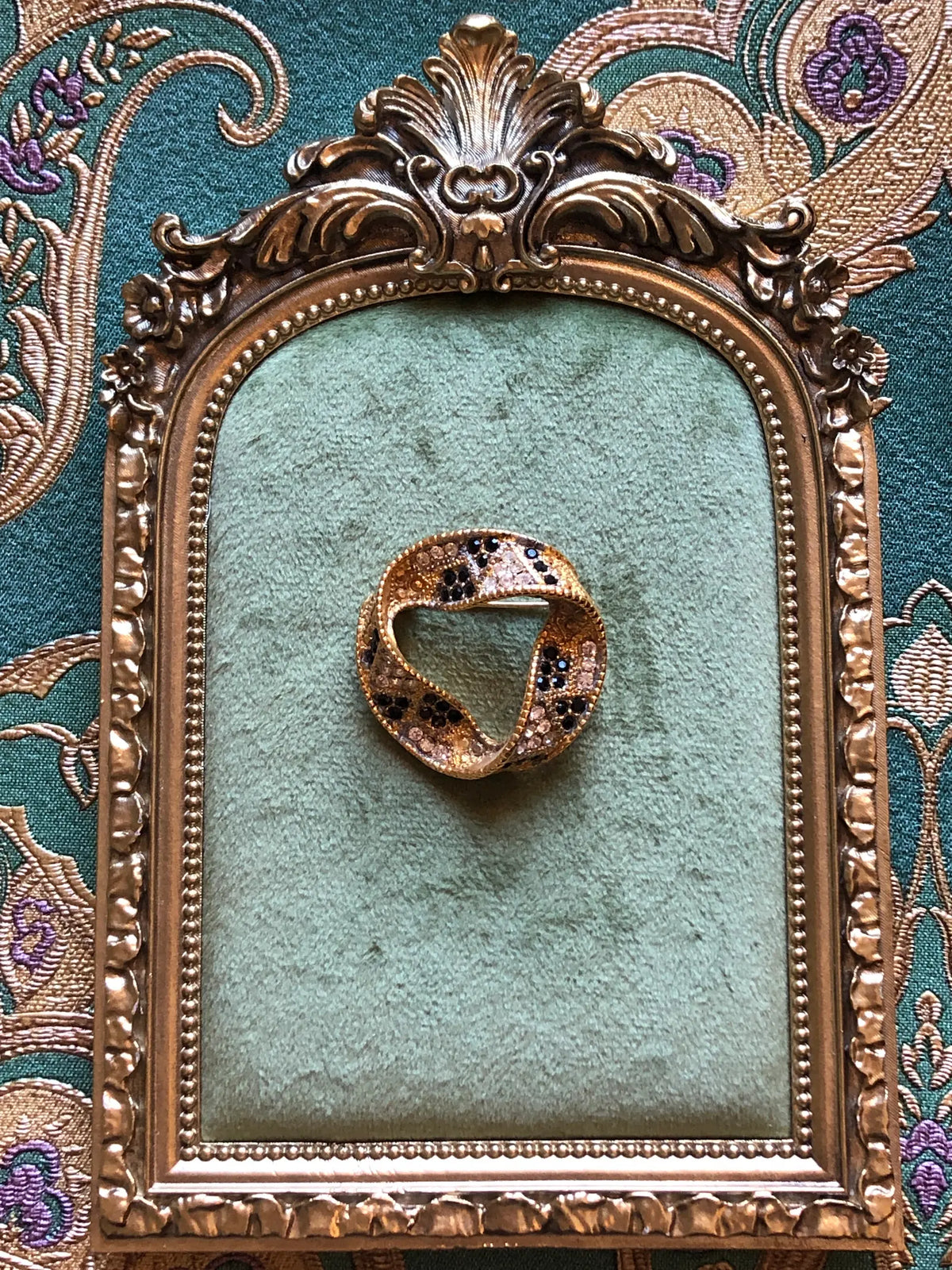 Broochella Men's Ottavia Vintage Black & Crystal Swirl Brooch featuring black and clear crystals set into a sculptural gold-tone circular design. Pictured on a green velvet tray with an ornate gold frame. The background is green and gold damask wallpaper. 