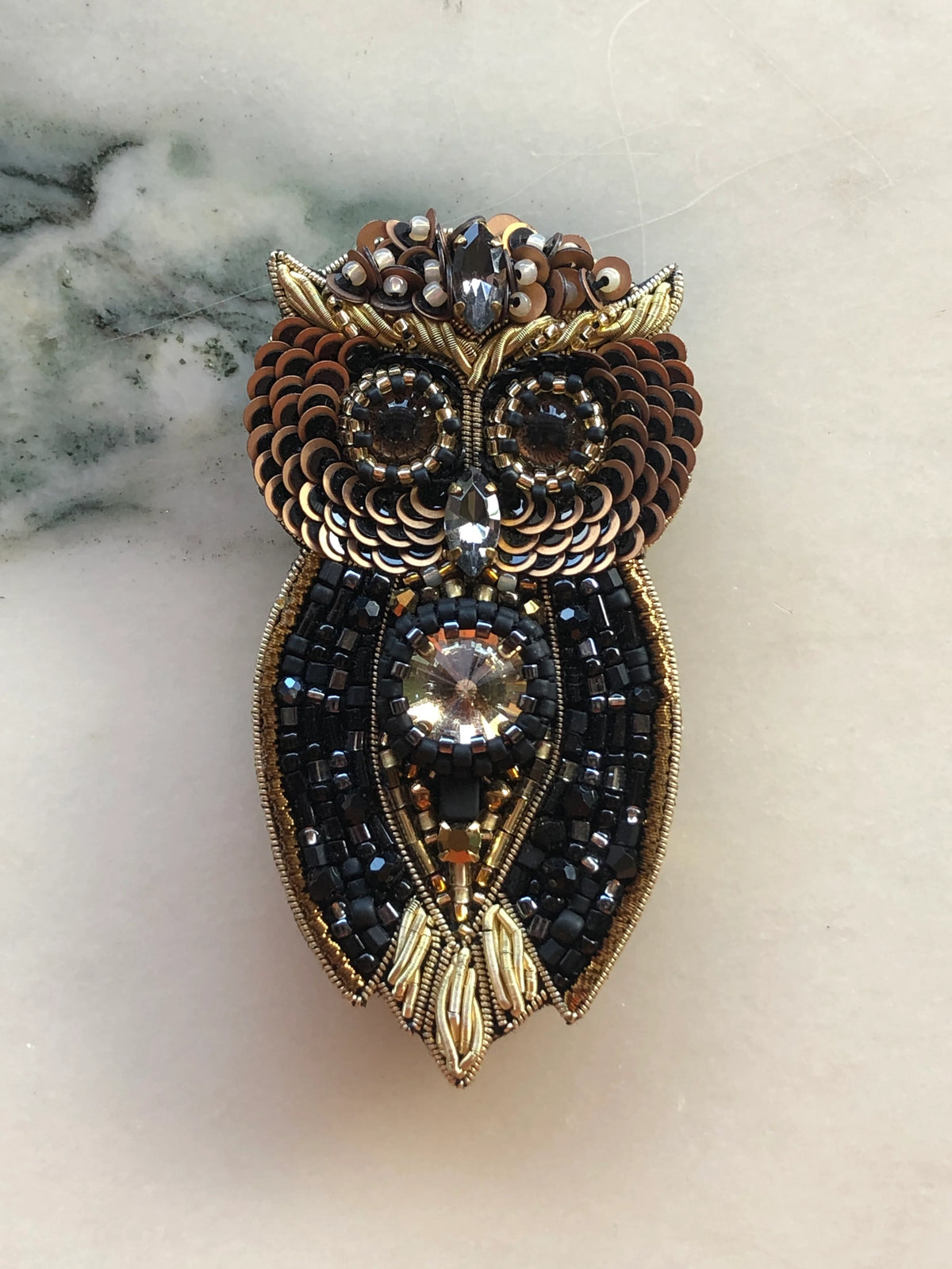Broochella Men’s Owl Brooch featuring intricate black and gold beadwork, bronze sequins and faceted crystal embellishments, photographed on a white marble background with grey-green veining.