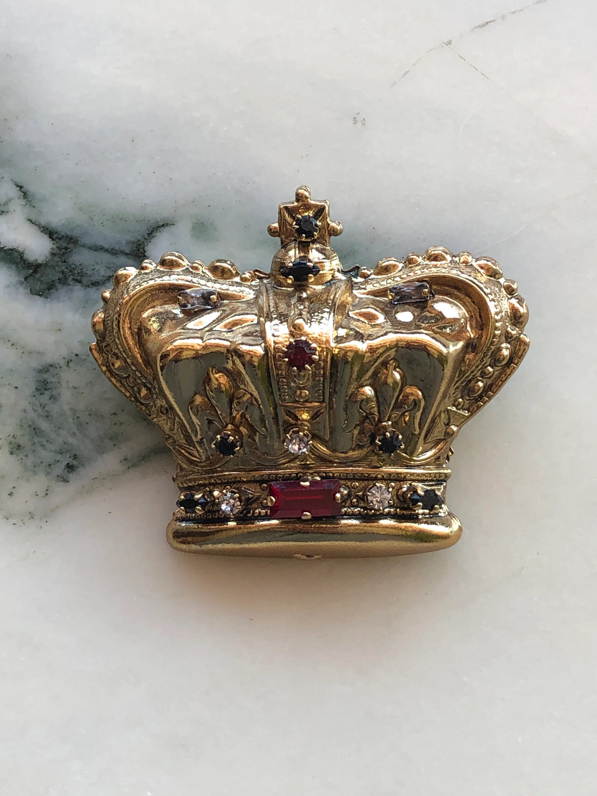 Broochella Men’s Regina Crown Brooch, an ornate gold crown design embellished with red, black and clear crystal stones, photographed on white marble.