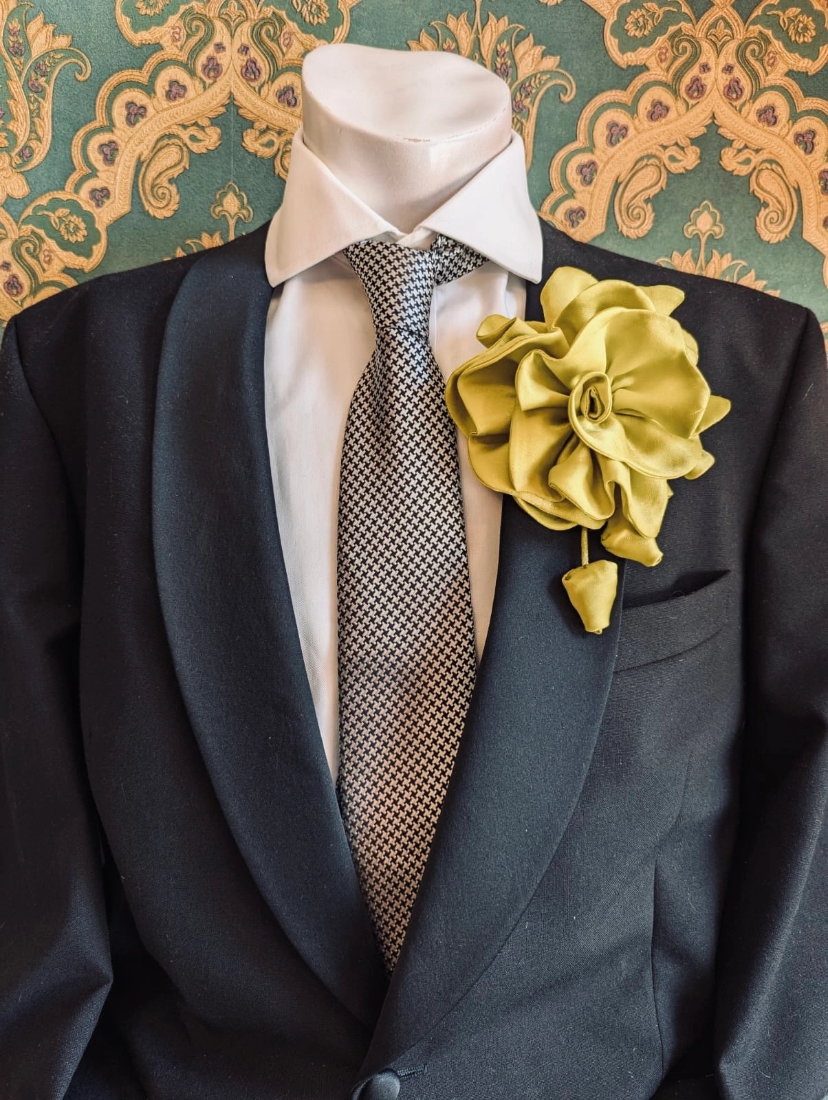 Broochella men's rosetta handmade, designer, satin flower brooch in chartreuse. Pinned on a men's mannequin wearing a black suit and a houndstooth tie. The background is green and gold damask wallpaper. 