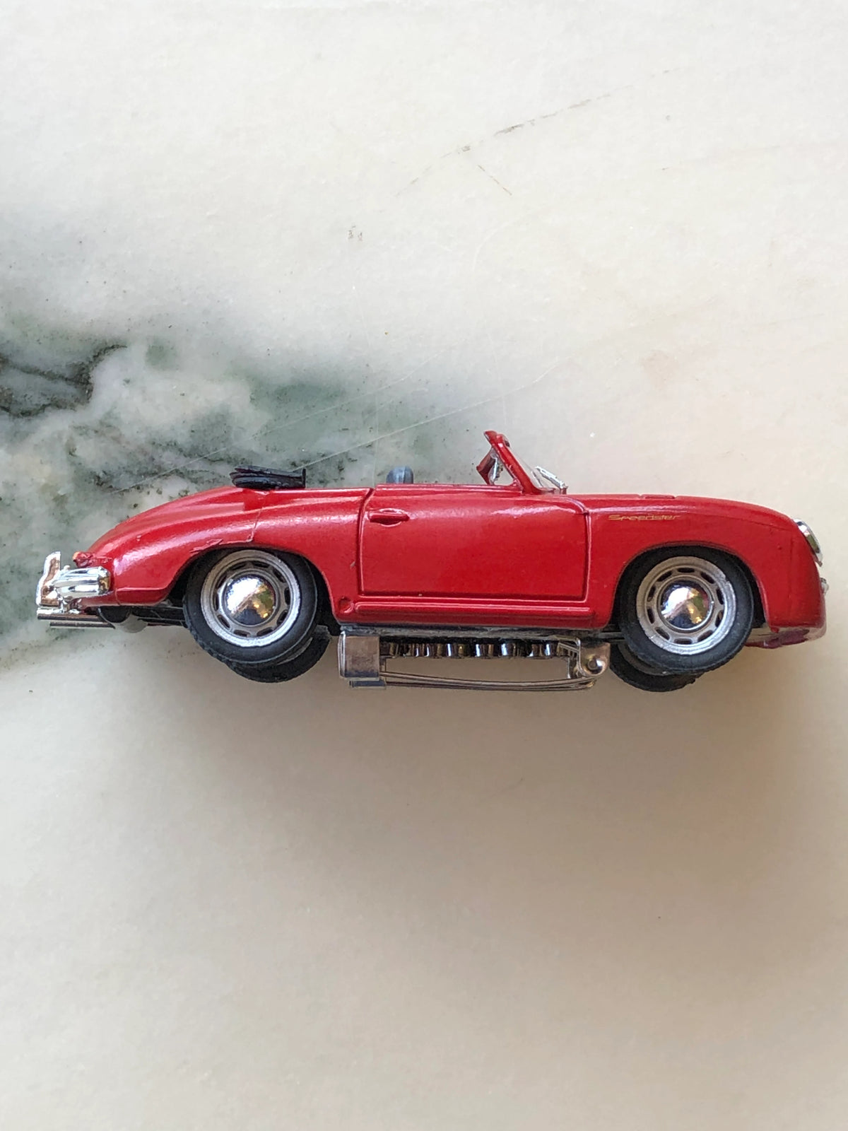 Broochella Men's Rosso Cabrio No.356 Vintage Convertible Car Brooch in side profile, featuring a classic red Speedster design, chrome hubcaps, black tyres, folded convertible roof and silver brooch pin fastening underneath.