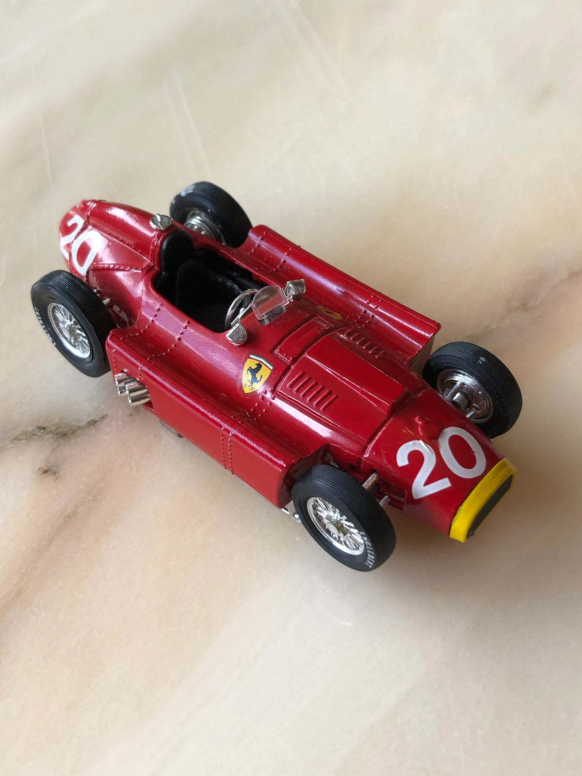 Broochella Men's Rosso Classico No.20 Ferrari Racing Car Brooch, featuring a vintage red open-cockpit race car with white number 20 decals, Ferrari shield emblems, silver spoked wheels and yellow nose detailing, photographed from an elevated angle.