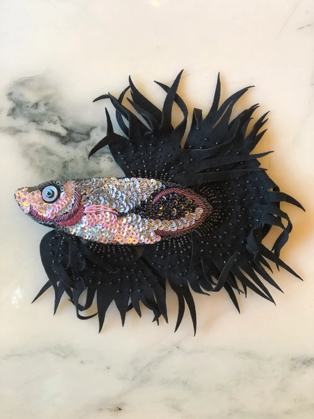 Broochella Men’s Siamese Fish Brooch, a statement handmade designer brooch intricately embellished with pink, silver and black sequins and beads, with dramatic black fabric fins, photographed on white marble.