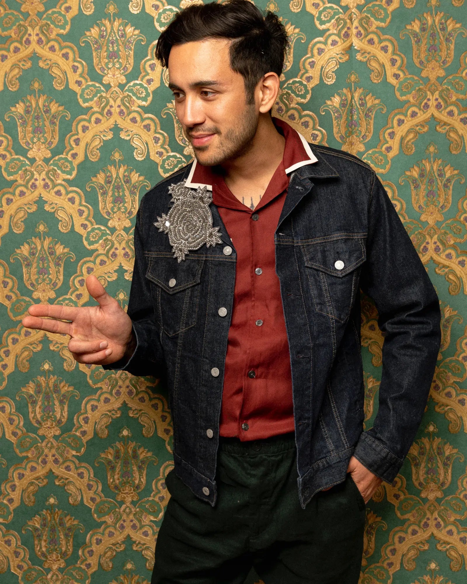 A man wearing a dark denim jacket with a silver embroidered patch on the chest over a rust red collared shirt and dark trousers, posing in front of green and gold damask wallpaper. He is wearing the Broochella Men's Stella silver hand beaded brooch. 