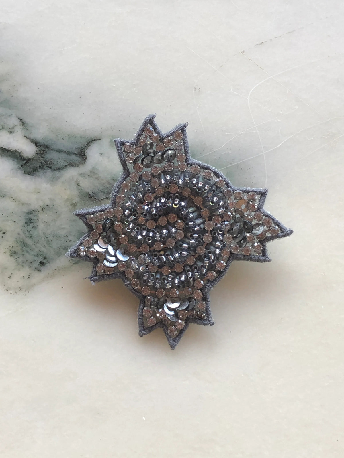 Broochella Men’s Stella small hand-beaded brooch pin, intricately embellished with silver and pewter beads, sequins and crystals in a sculptural starburst design, photographed on white marble.