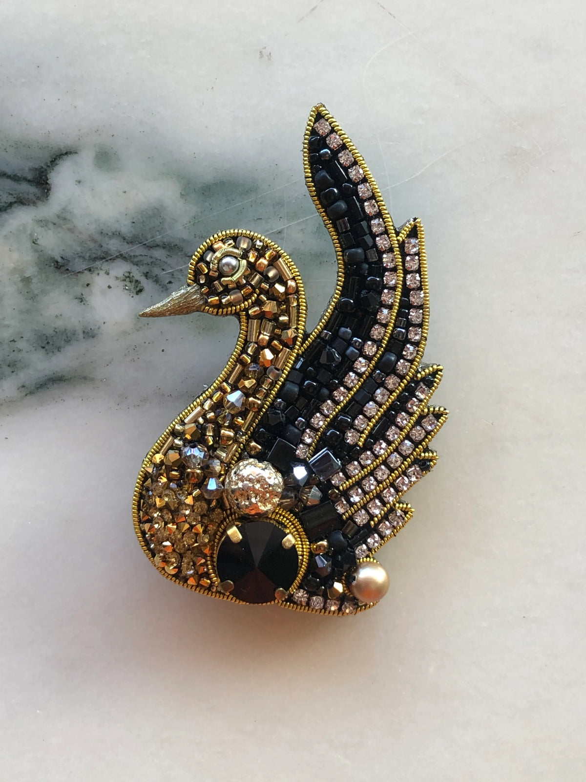Broochella Men’s Swan Brooch featuring intricate black and gold beadwork, sparkling crystal detailing and pearl accents, photographed on a white marble background with grey-green veining.