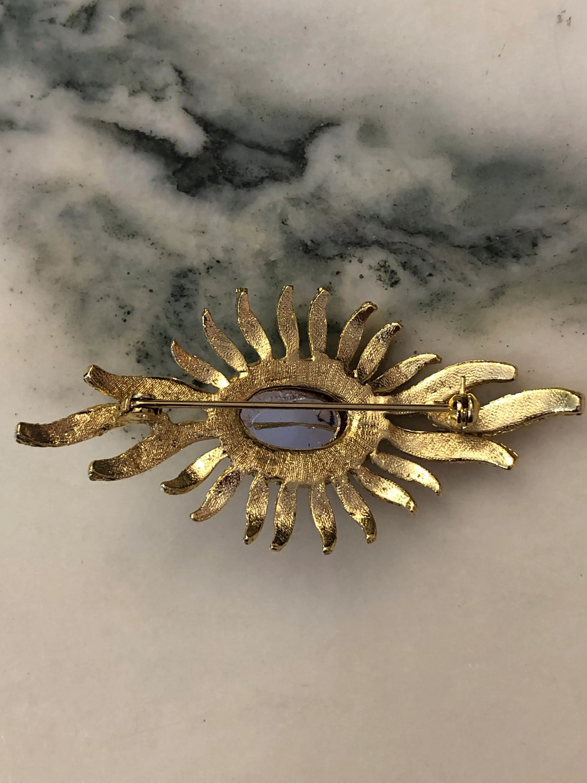The back view of Broochella Men's Valentina Vintage Purple Sunburst Brooch featuring a deep-purple oval cabochon surrounded by gold-tone crystal-set rays on a cream and grey marble background.