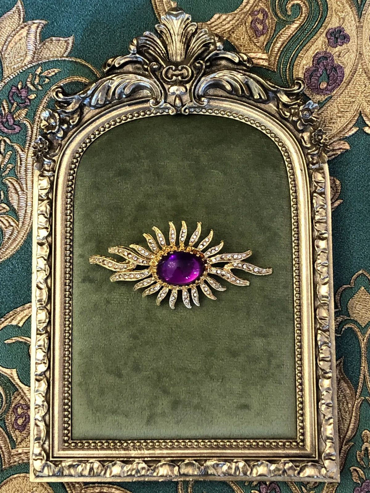 Broochella Men's Valentina Vintage Purple Sunburst Brooch featuring a deep-purple oval cabochon surrounded by gold-tone crystal-set rays. It is displayed on a green velvet tray with an ornate gold frame.