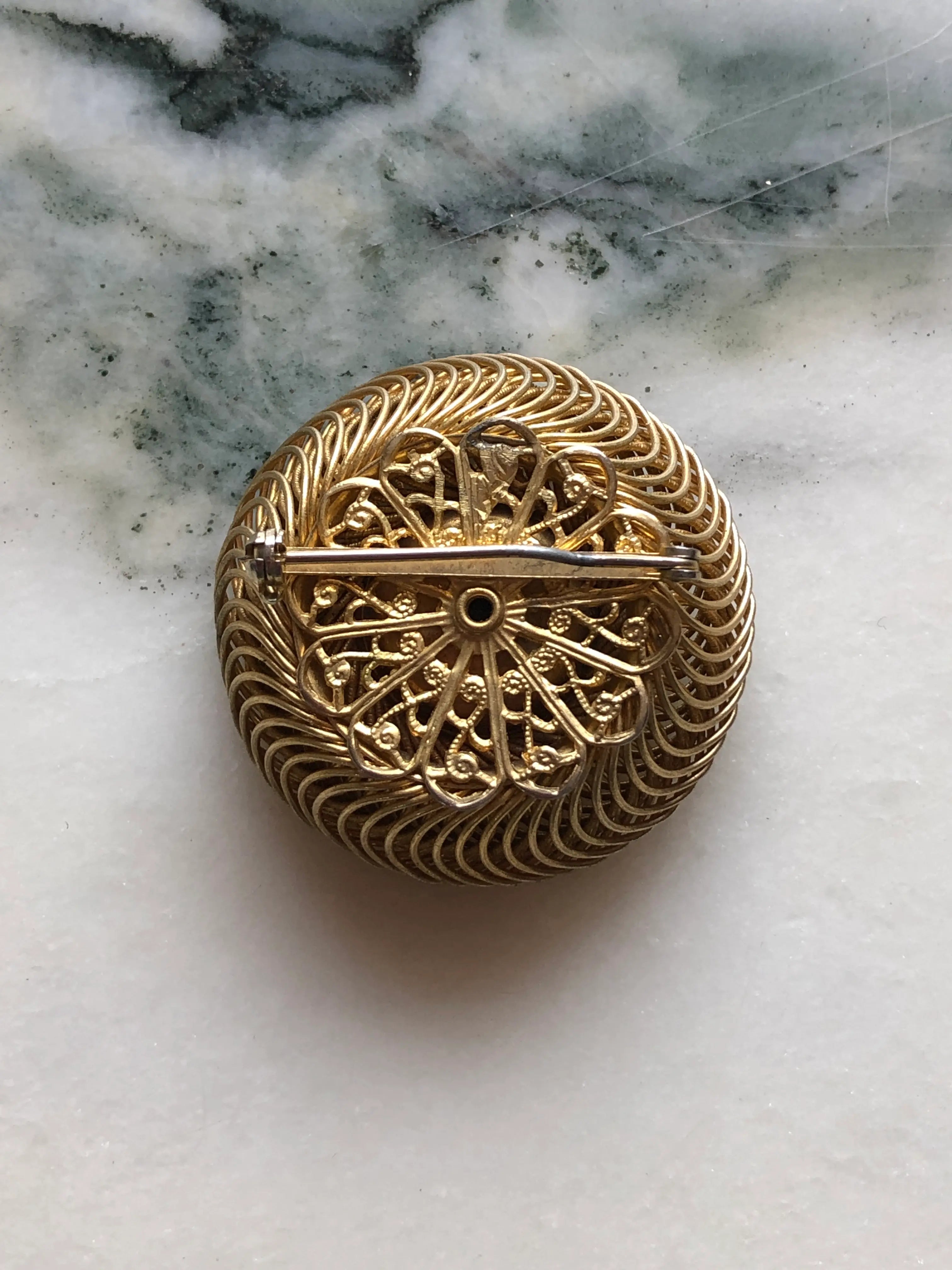 The back view of Broochella Midnight Halo vintage circular brooch with a glossy black domed cabochon and intricate spiral gold-tone wirework, measuring approximately 4 cm. Photographed on white and grey marble.