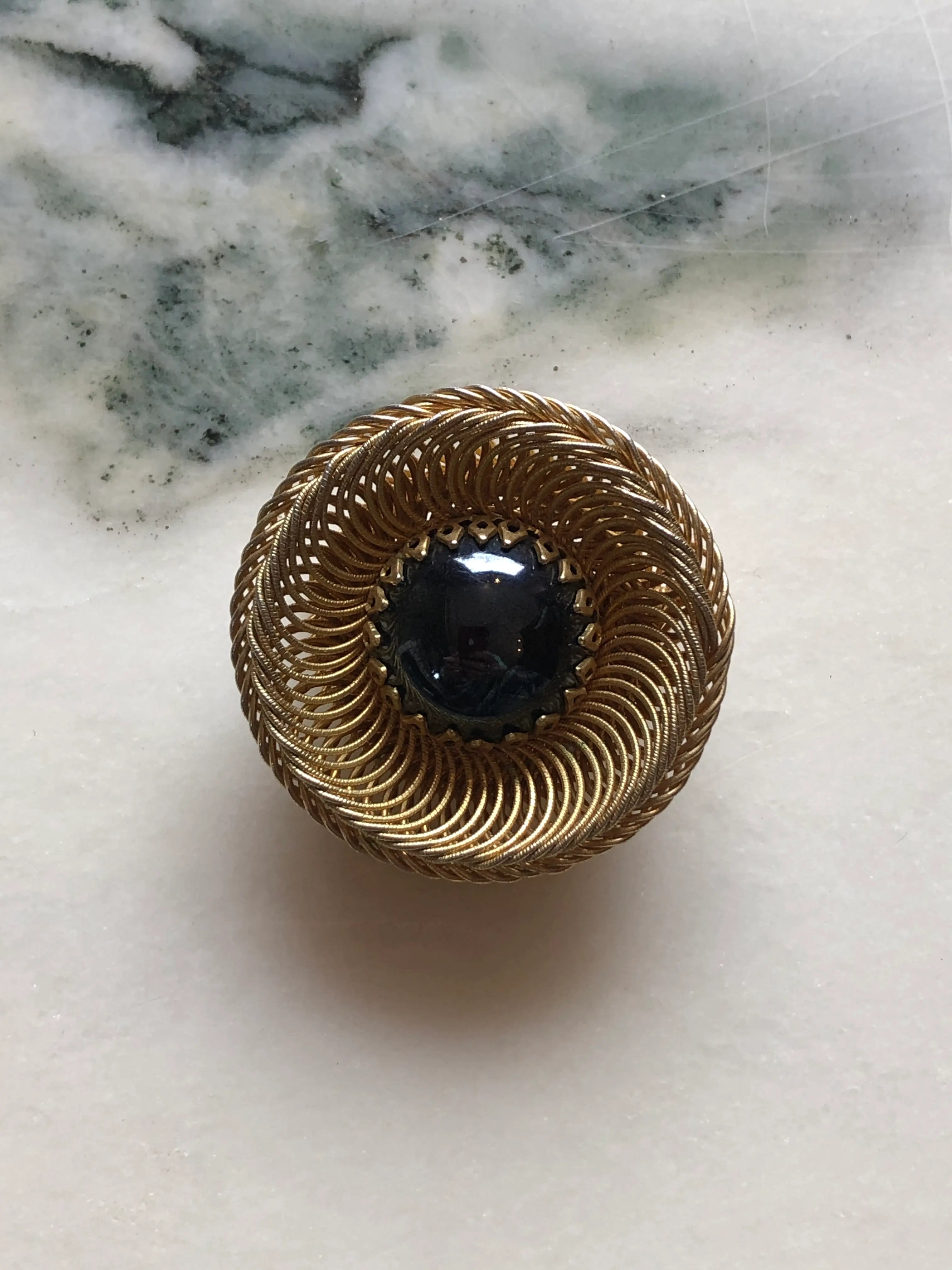 Broochella Midnight Halo vintage circular brooch with a glossy black domed cabochon and intricate spiral gold-tone wirework, measuring approximately 4 cm. Photographed on white and grey marble.