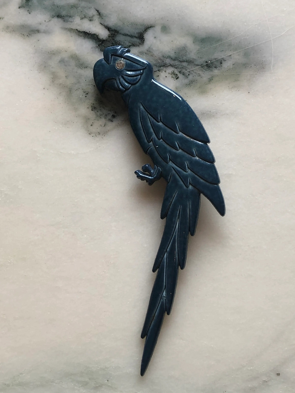 Broochella Midnight Macaw vintage Italian parrot brooch in deep blue with a clear crystal eye, sculpted wings and a long feathered tail, measuring approximately 13.5 × 5 cm. Photographed on white and grey marble.
