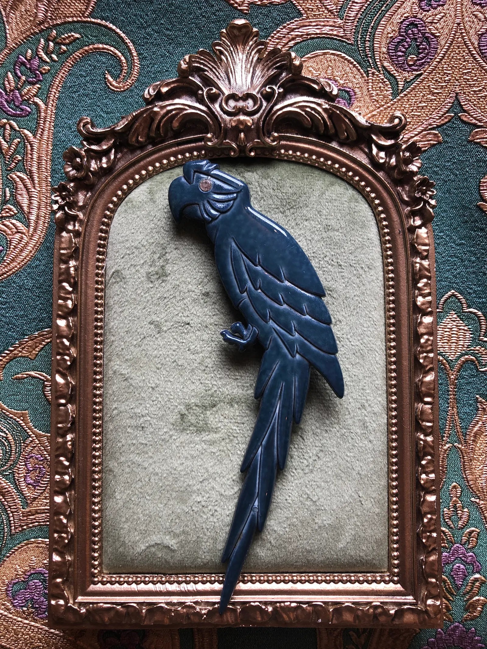 Broochella Midnight Macaw vintage Italian parrot brooch in deep blue with a clear crystal eye, sculpted wings and a long feathered tail, measuring approximately 13.5 × 5 cm. Photographed on a green velvet tray with an ornate gold frame. The background is green and gold damask wallpaper.