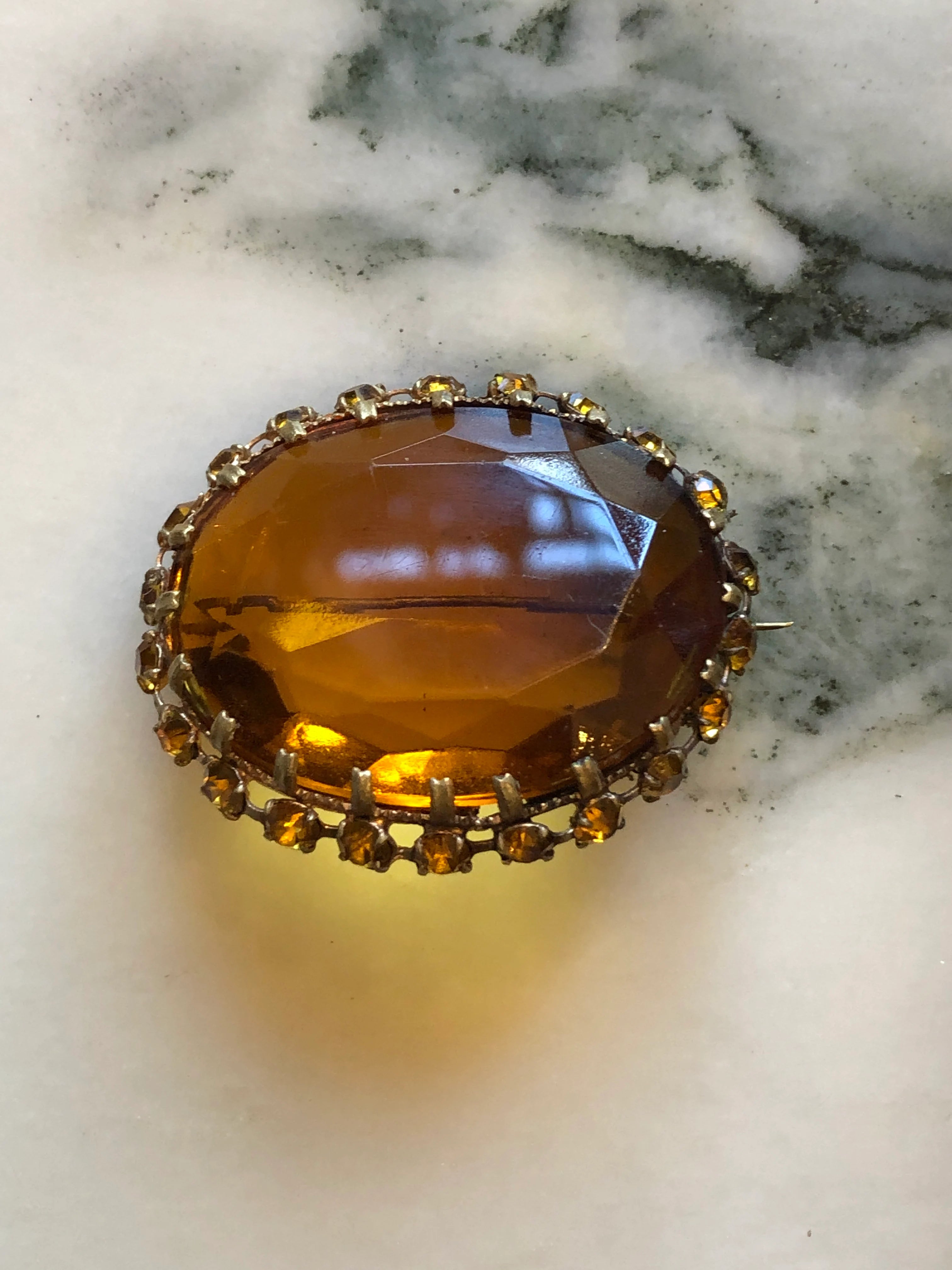 Broochella Miele Imperiale vintage gold-tone brooch with an oversized oval amber crystal and coordinating crystal halo. The background is white marble.
