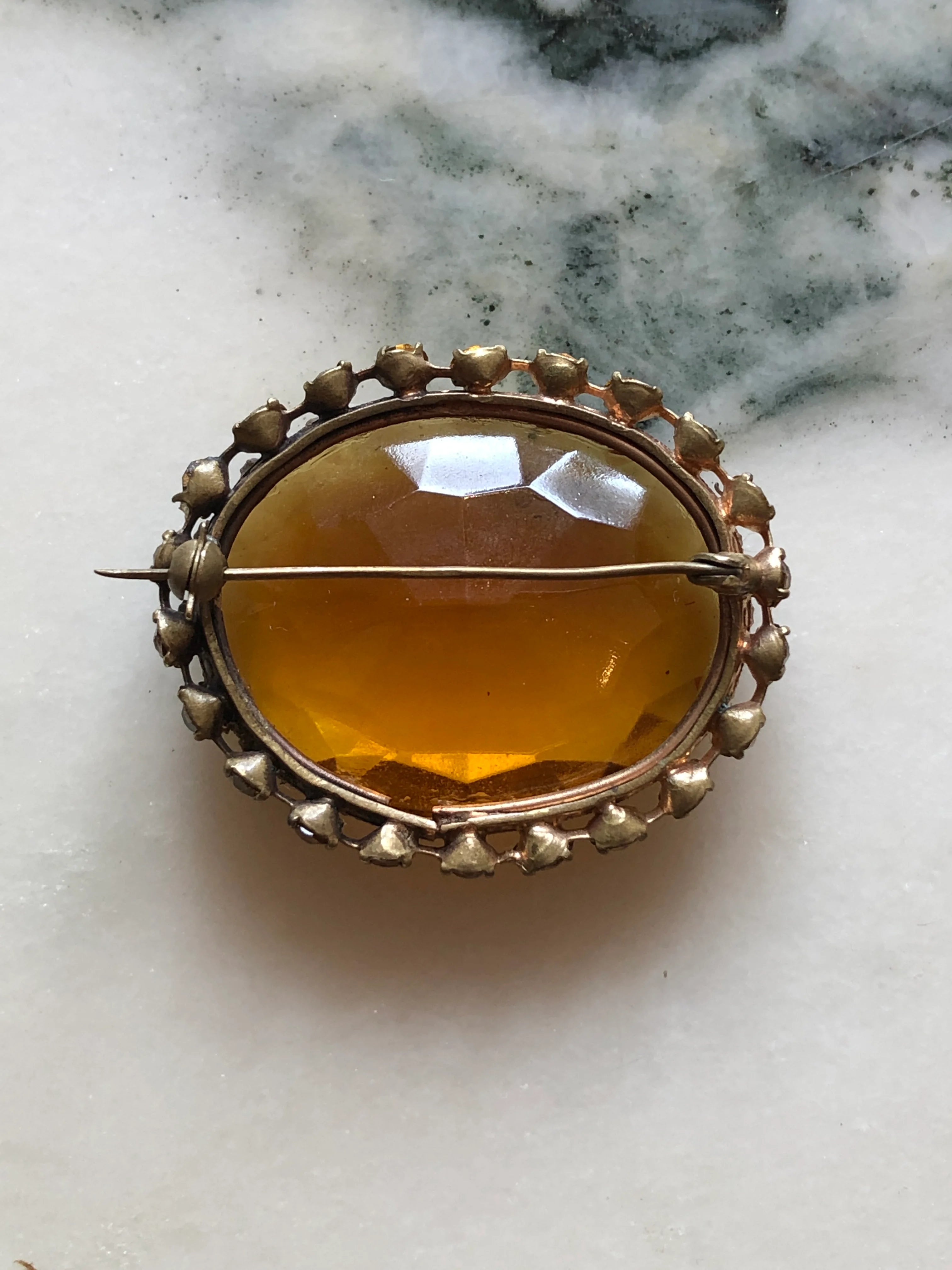 The back view of Broochella Miele Imperiale vintage gold-tone brooch with an oversized oval amber crystal and coordinating crystal halo. The background is white marble.
