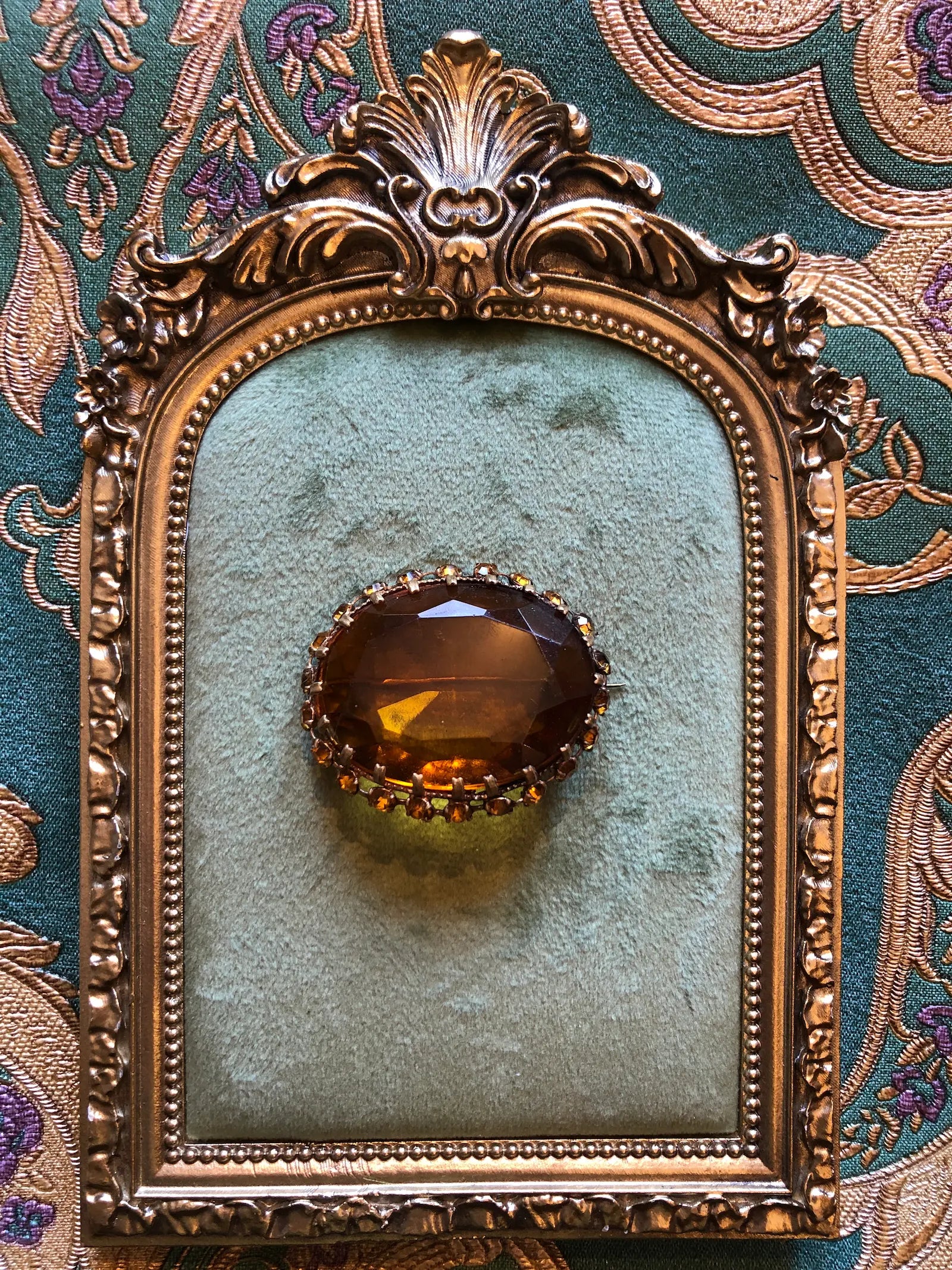 An ornate Broochella Miele Imeperiale Vintage oval brooch featuring a large faceted amber-brown center stone surrounded by small red and gold stones, displayed on a light green velvet insert within an antique gold frame, set against a patterned teal and gold fabric background.