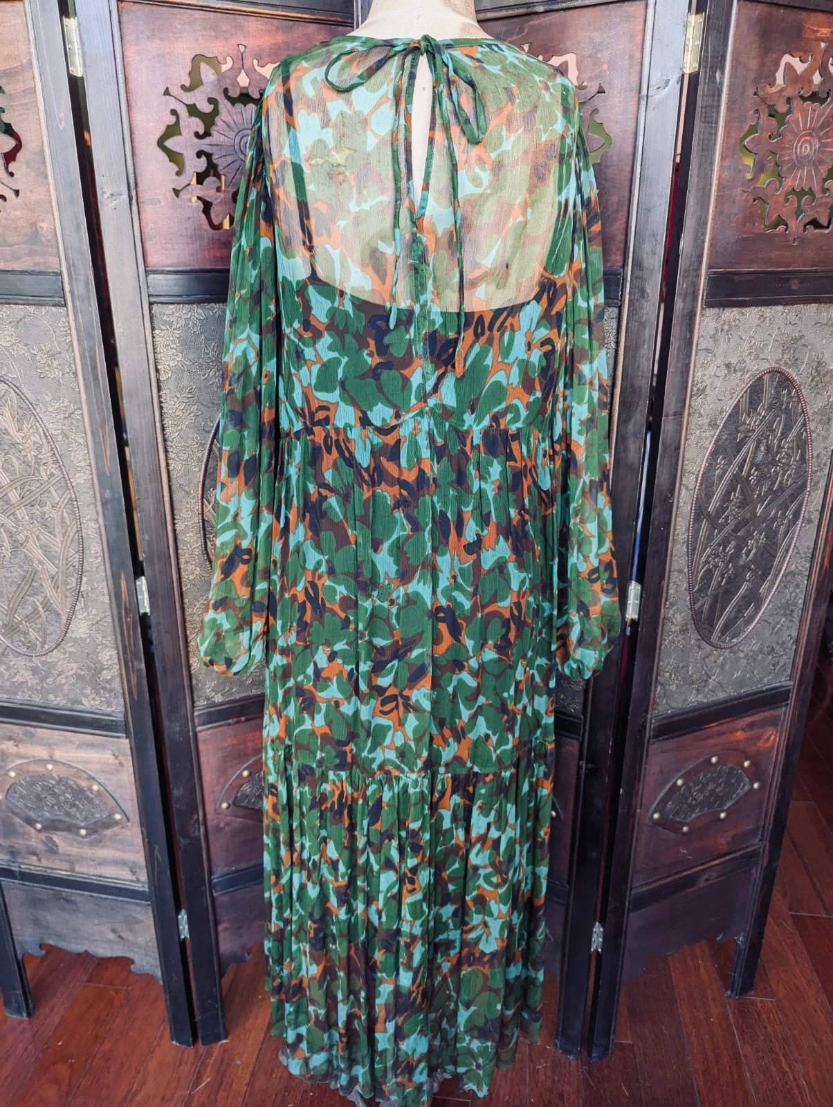 Broochella MLM Green,  blue, rust and black abstract floral, sheer, tiered, midi Dress with Slip – Size Small. Pictured on a mannequin. The background is an ornate, dark wood room divider. 