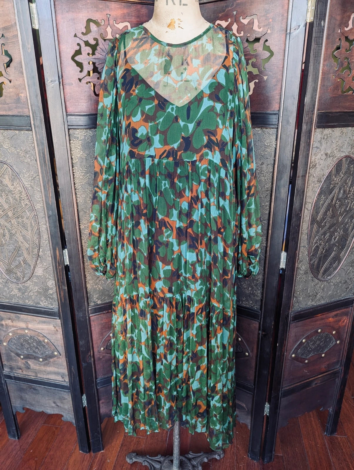 Broochella MLM Green,  blue, rust and black abstract floral, sheer, tiered, midi Dress with Slip – Size Small. Pictured on a mannequin. The background is an ornate, dark wood room divider. 