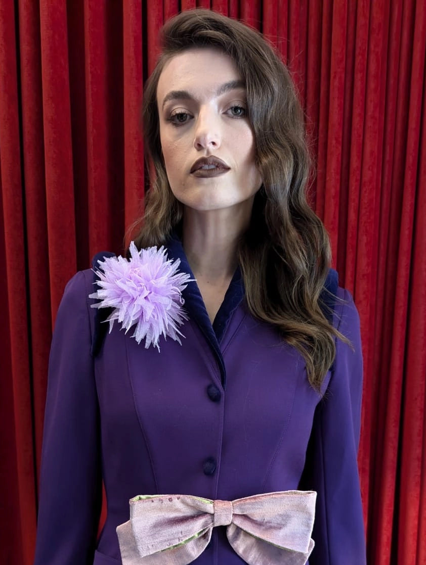 A model stands in front of a red velvet curtain wearing a purple long-sleeve jacket with a large light pink bow at the waist and the Broochella monello lilac urchin brooch