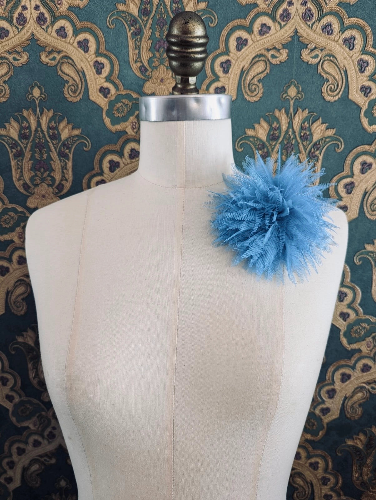 Broochella monello handmade tulle urchin brooch in blue. The brooch is pinned on a mannequin. The background is green and gold damask wallpaper.