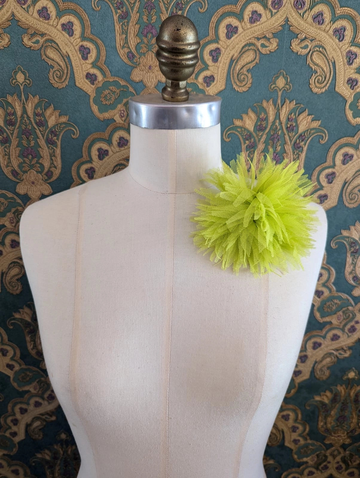 Broochella monello handmade tulle urchin brooch in chartreuse. The brooch is pinned on a mannequin. The background is green and gold damask wallpaper.