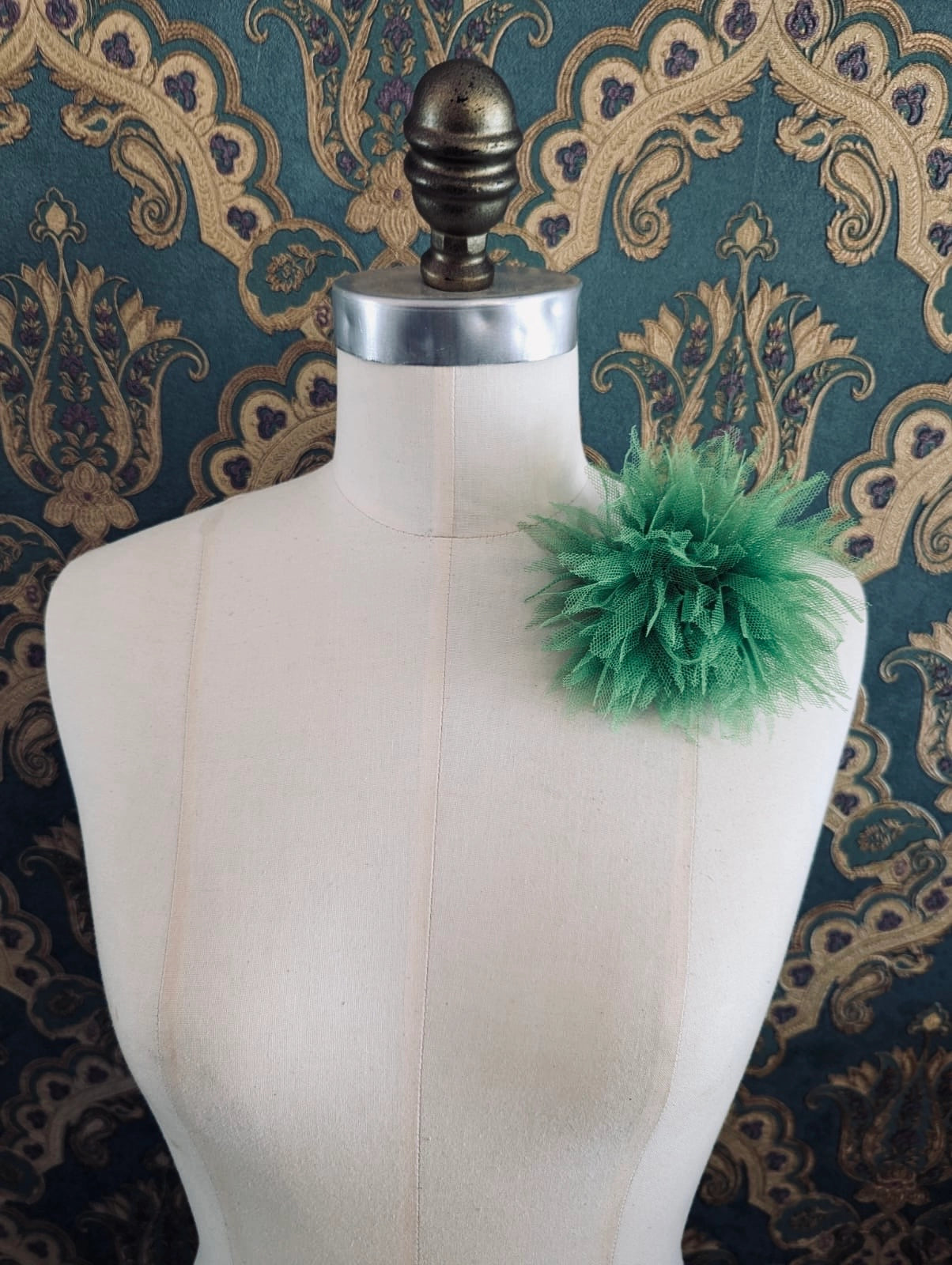 Broochella monello handmade tulle urchin brooch in green. The brooch is pinned on a mannequin. The background is green and gold damask wallpaper.