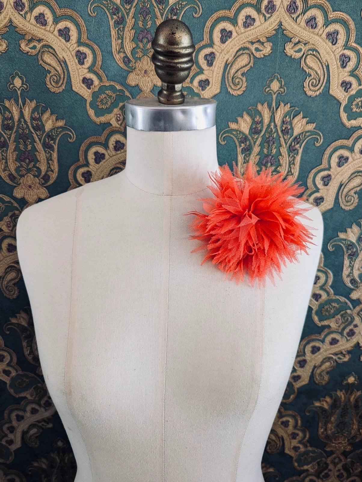 Broochella monello handmade tulle urchin brooch in orange. The brooch is pinned on a mannequin. The background is green and gold damask wallpaper.