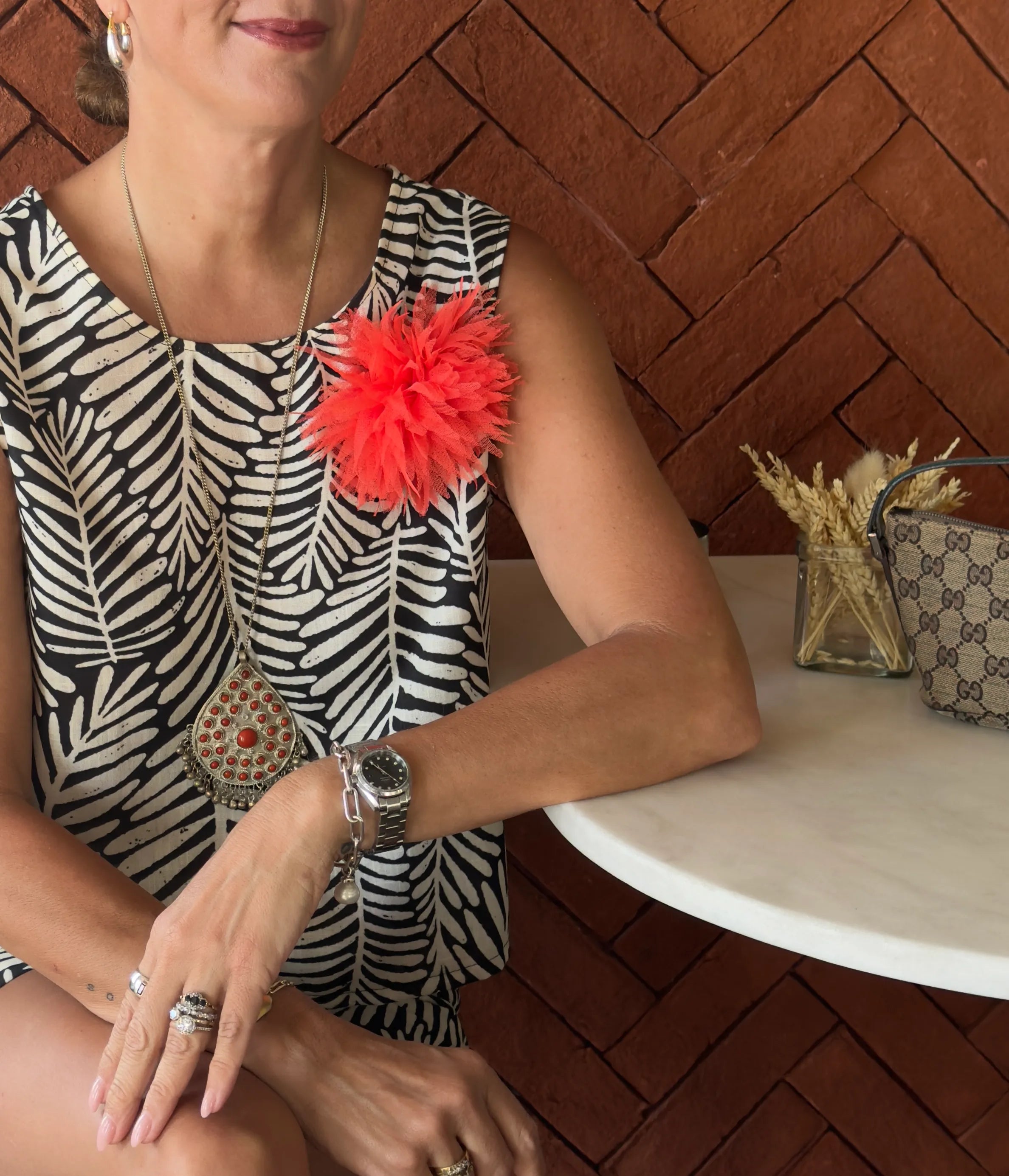 A seated woman in a matching black and white geometric top and shorts. She is sitting at a table at a cafe. She is wearing the Broochella Monello urchin brooch in orange. The background is a herringbone brick wall. 