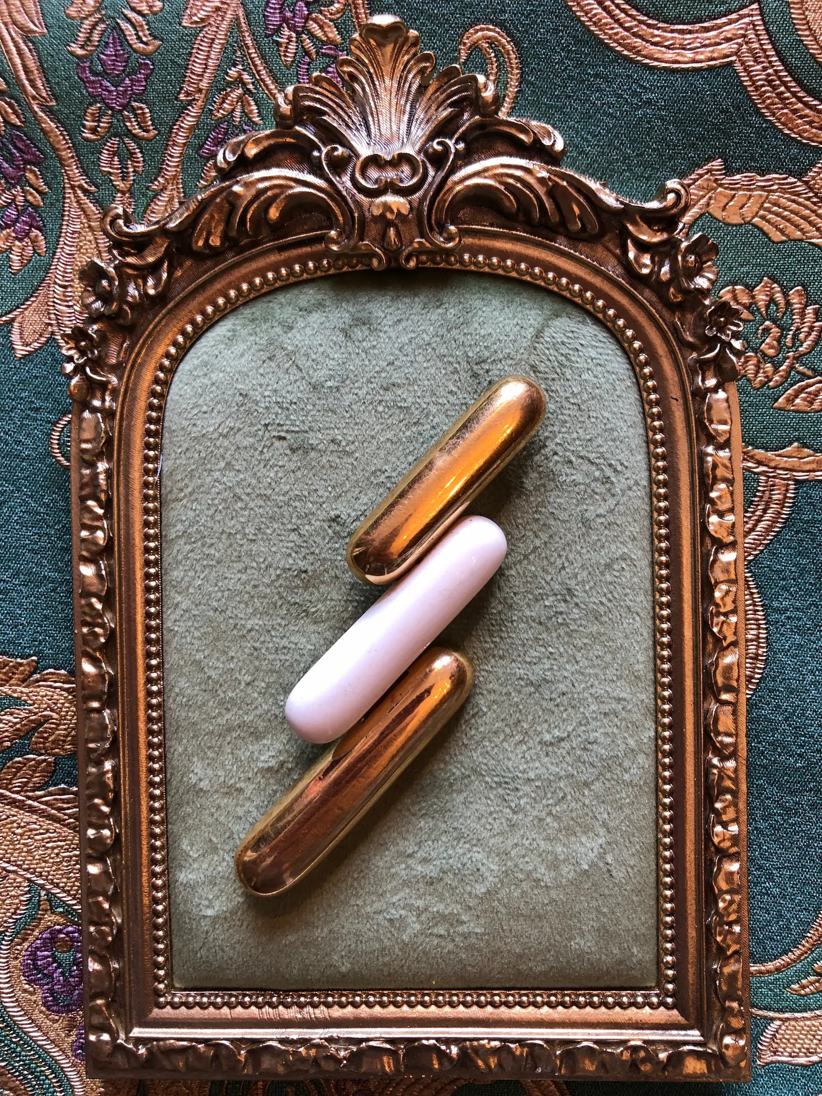 Broochella Monet Vintage Directives brooch with three elongated gold-tone and ivory enamel geometric bars. It is Arranged diagonally inside an ornate vintage-style gold frame on a textured green surface with a patterned fabric background.