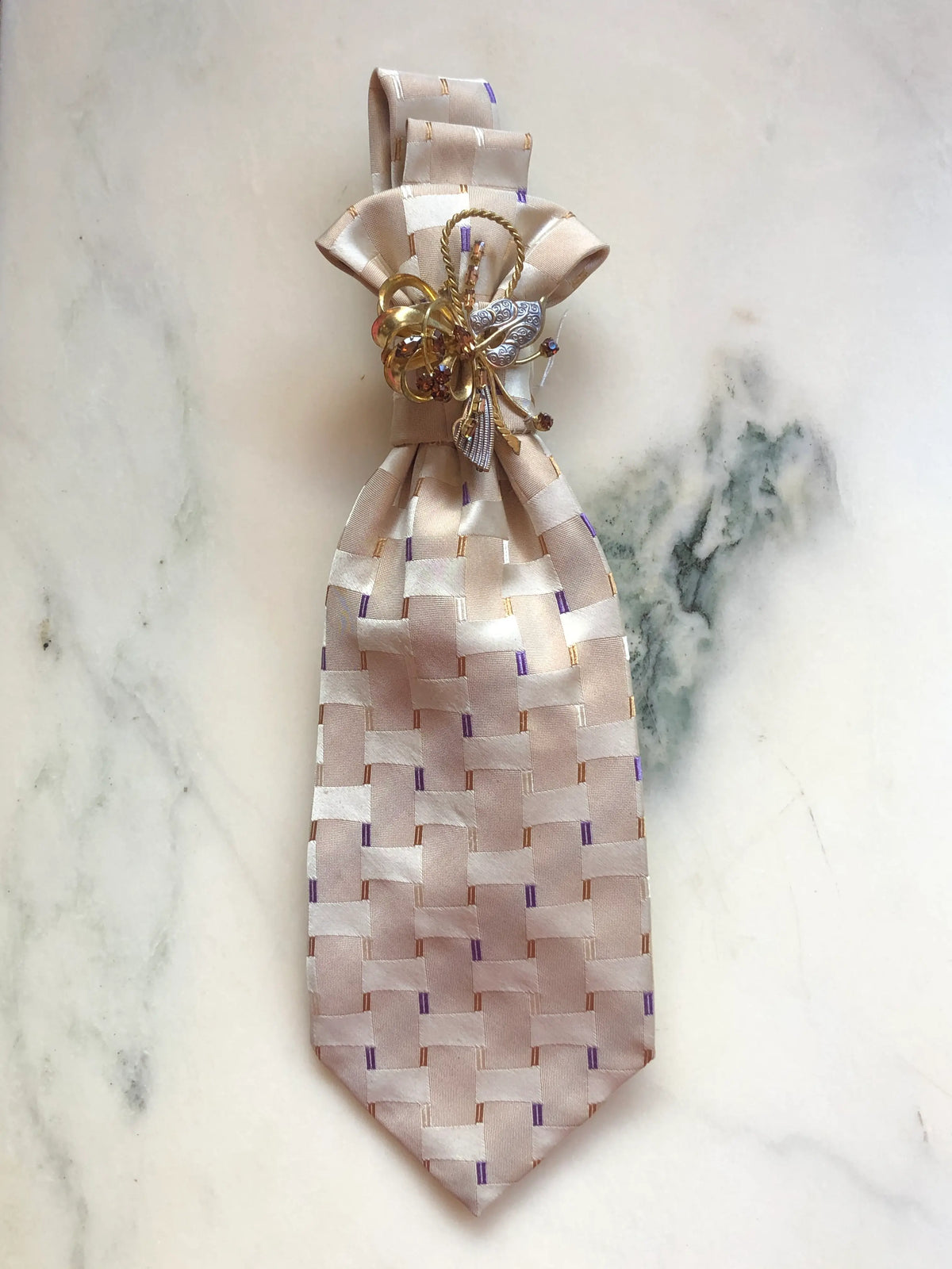 Broochella Montecristo Cravatta Tie Brooch Pin #22, a handmade designer statement brooch created from an upcycled vintage ivory and blush woven silk tie with subtle purple and gold accents, finished with an ornate mixed-metal vintage-style floral embellishment, photographed on white marble.
