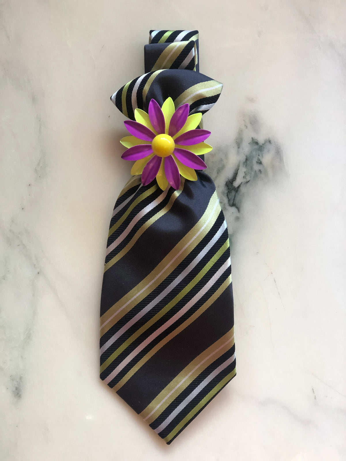 Broochella Montecristo Cravatta Tie Brooch Pin #24, a handmade designer statement brooch crafted from an upcycled vintage black striped tie with cream, lime detailing, finished with a bold purple and yellow floral embellishment and photographed on white marble.