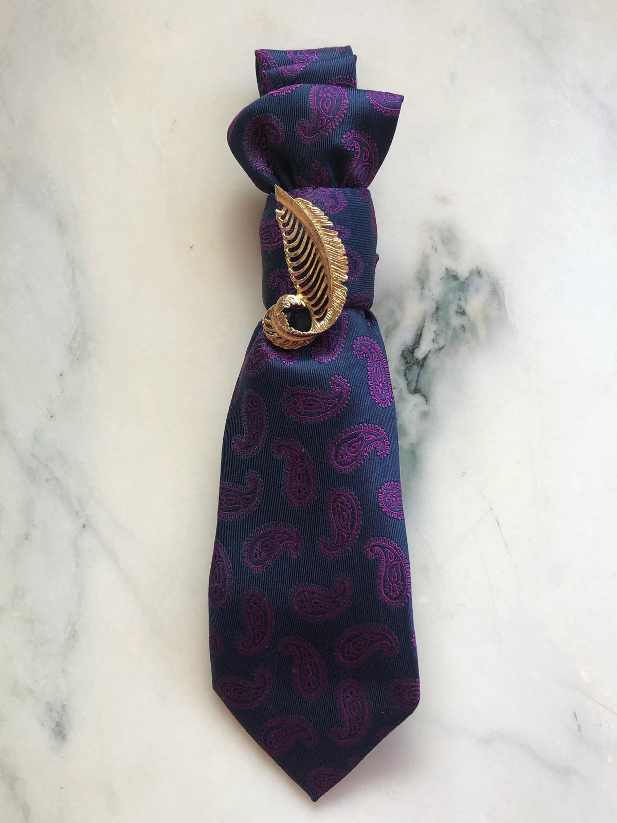 Broochella Montecristo Cravatta Tie Brooch Pin #9, a handmade designer statement brooch crafted from an upcycled vintage navy tie with magenta paisley pattern, gathered and finished with a sculptural gold-tone leaf brooch, photographed on white marble.