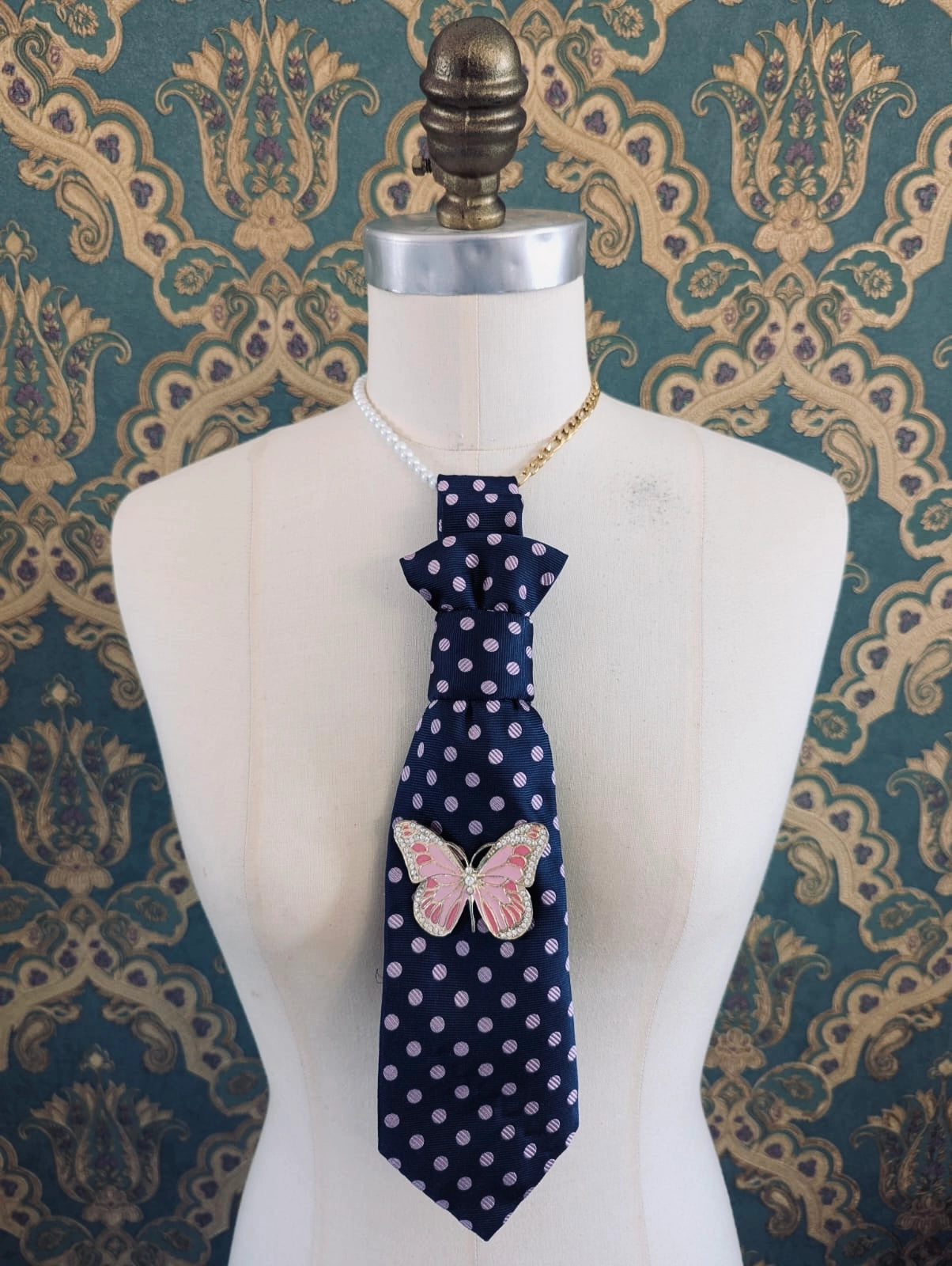 Broochella Montecristo cravatta vintage upcycled tie brooch in navy with pink polka dots. It has a vintage pink butterfly brooch pinned to the front. It is displayed on a mannequin. The background is green and gold damask wallpaper. 
