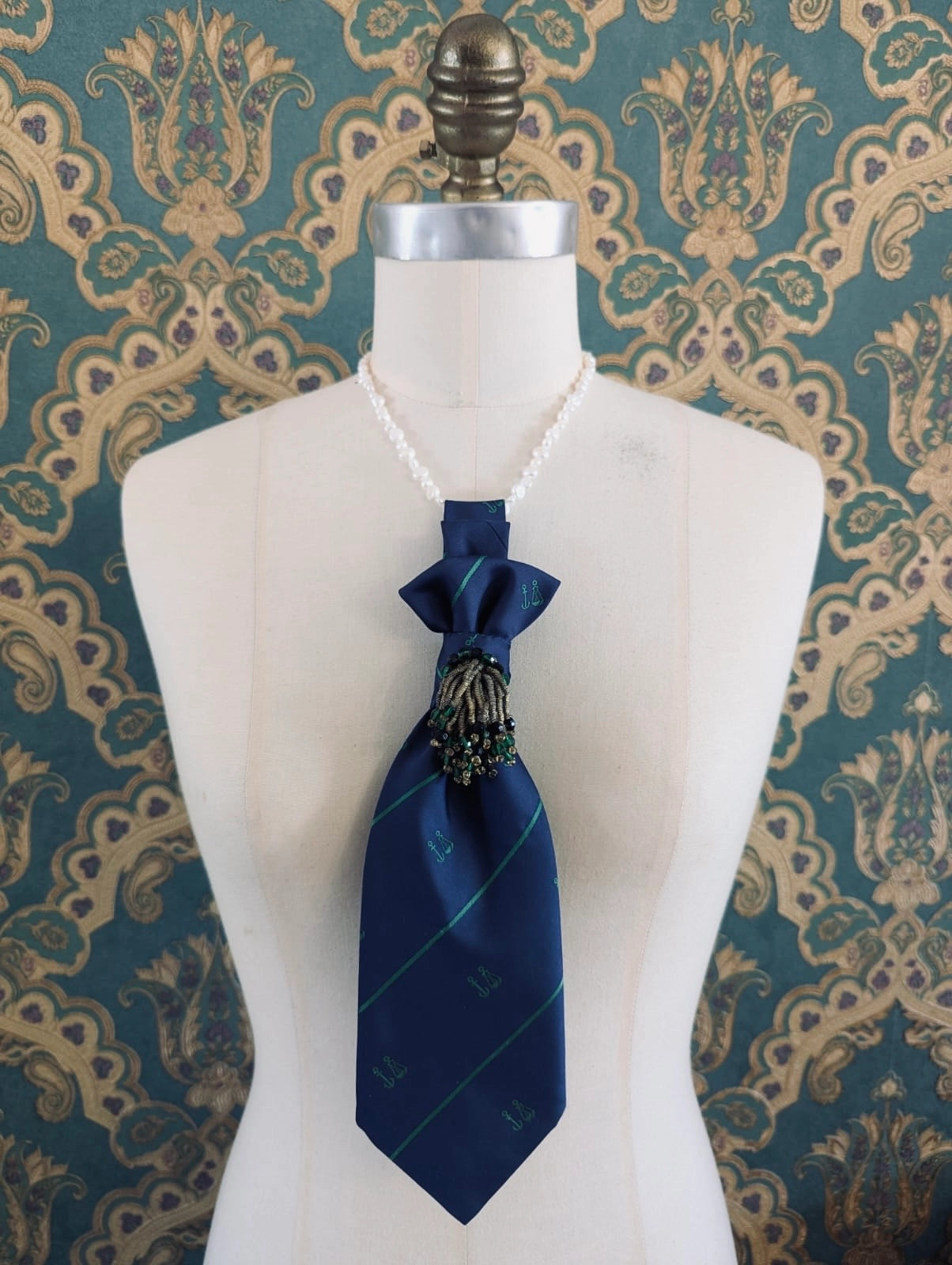 A Broochella Montecristo cravatta number 19 upcycled vintage tie brooch in navy and green stripe, with a vintage brooch embellishment. It is pinned on a cream-coloured dress form mannequin. The background is green and gold damask wallpaper. 